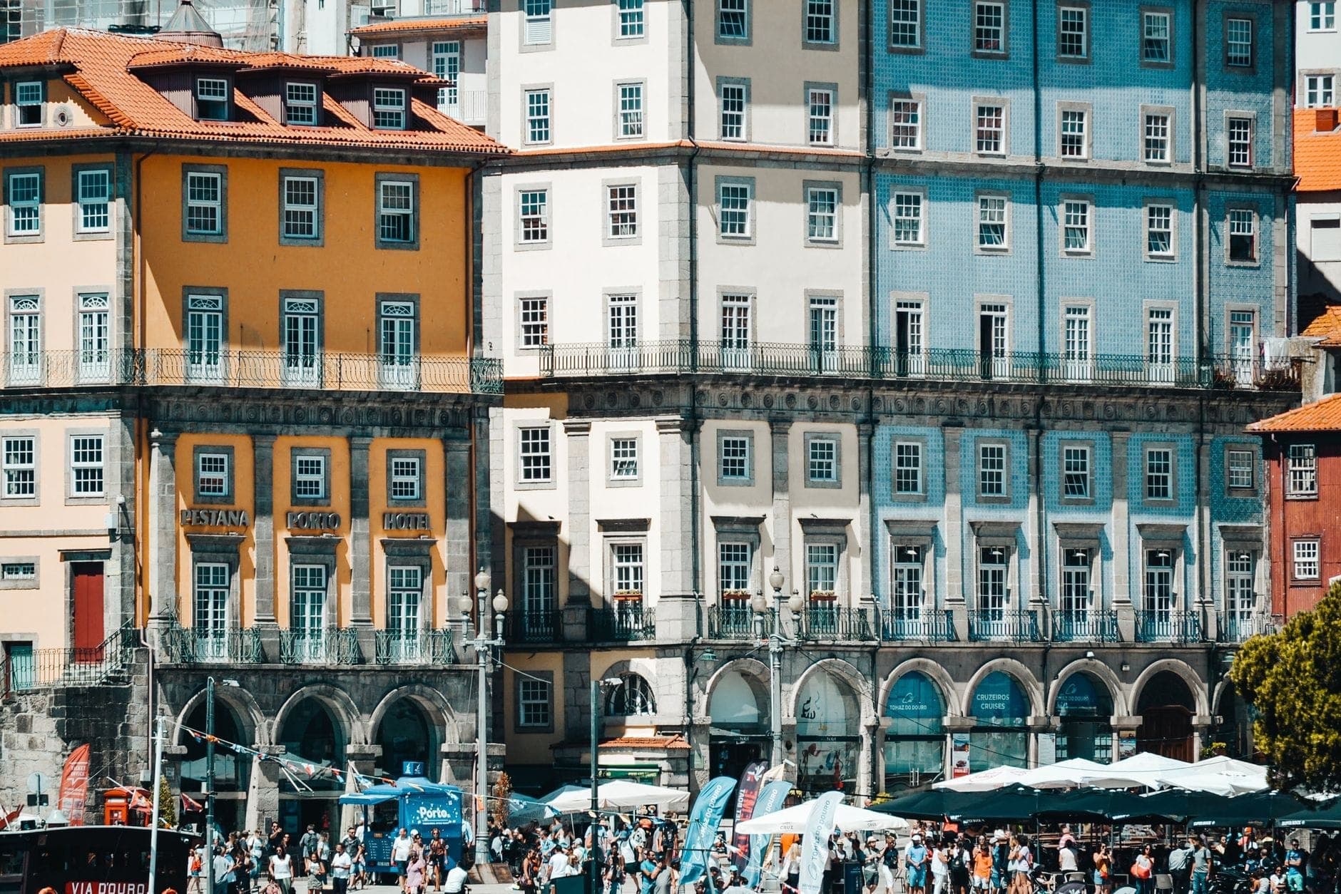 Multidão agitada de verão no histórico bairro da Ribeira no Porto, com edifícios antigos coloridos, cafés na rua e espaços ao ar livre lotados, ilustrando a época alta do turismo.