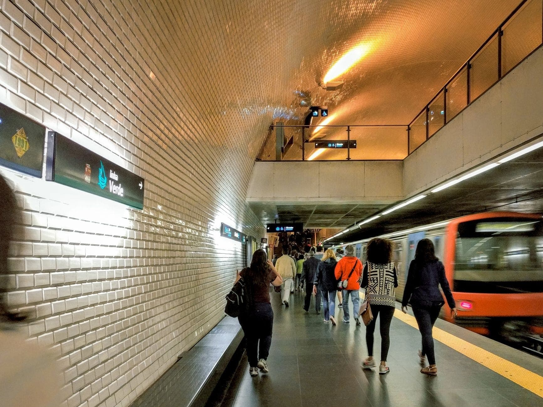 Passageiros caminhando ao longo de uma plataforma de metrô bem iluminada no Porto, com um trem de metrô chegando e sinalizações da estação visíveis na parede de azulejos.