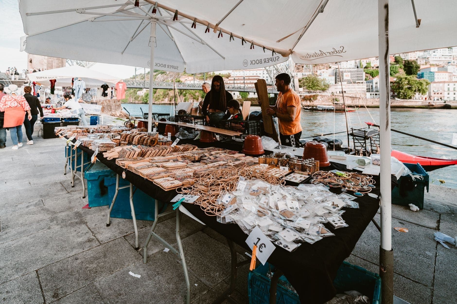 Freiluftmarkt am Flussufer in Porto mit Ständen, die lokales Kunsthandwerk und Lebensmittel unter weißen Schirmen verkaufen – im Hintergrund flanieren Besucher vor dem Fluss.