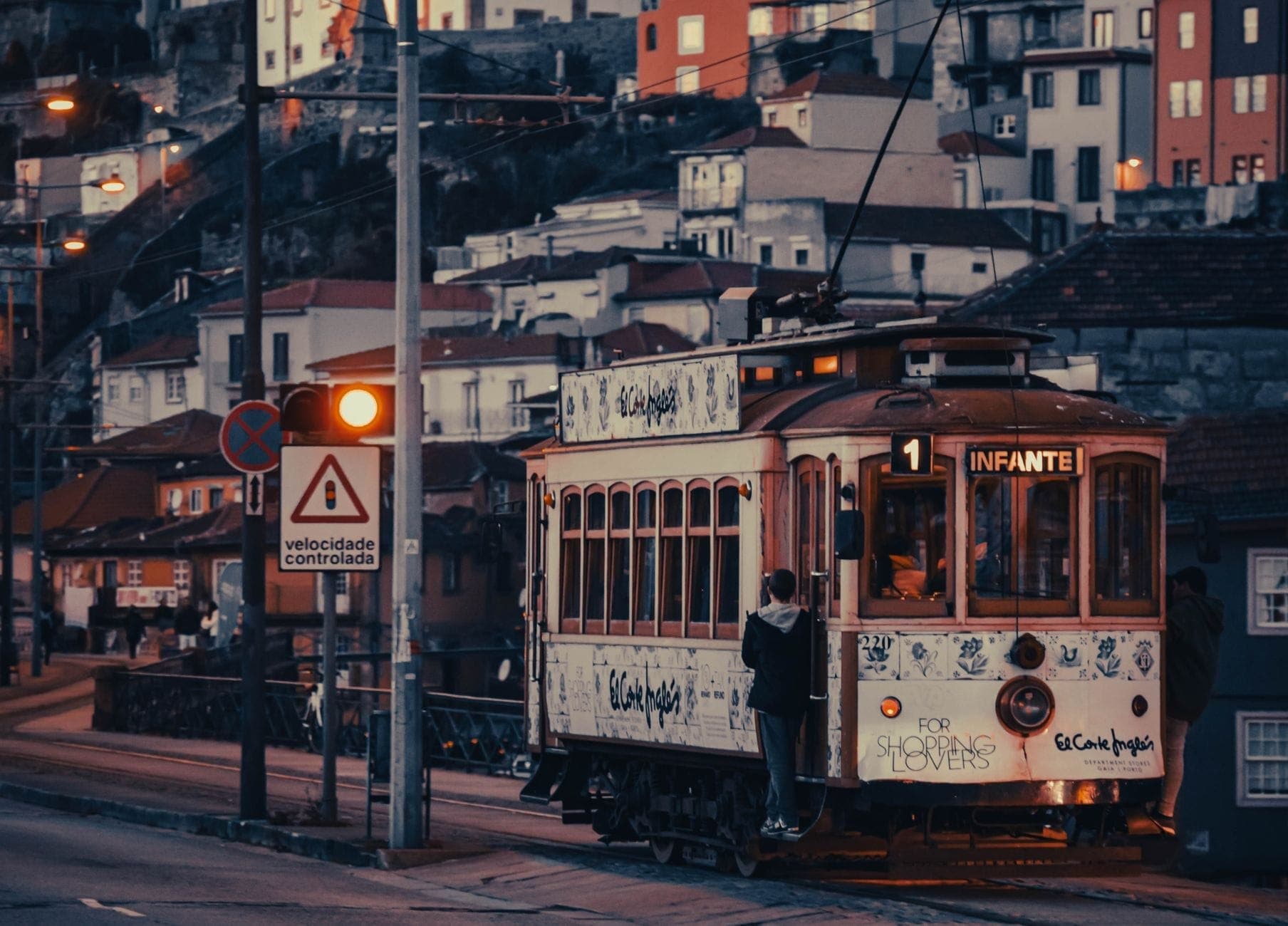 Historischer Tramwagen mit der Aufschrift Infante auf Portos Linie 1 fährt bei Abenddämmerung entlang des Douro, im Hintergrund bunte Häuser auf dem Hang.