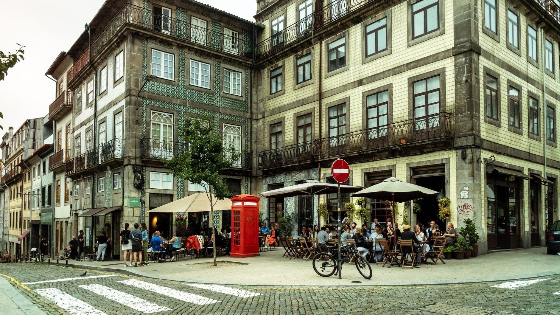 Widok na róg tętniącej życiem dzielnicy Porto z wyłożonymi kafelkami budynkami, ogródkami kawiarnianymi, jedzącymi ludźmi i czerwoną budką telefoniczną w stylu brytyjskim na brukowanej ulicy.