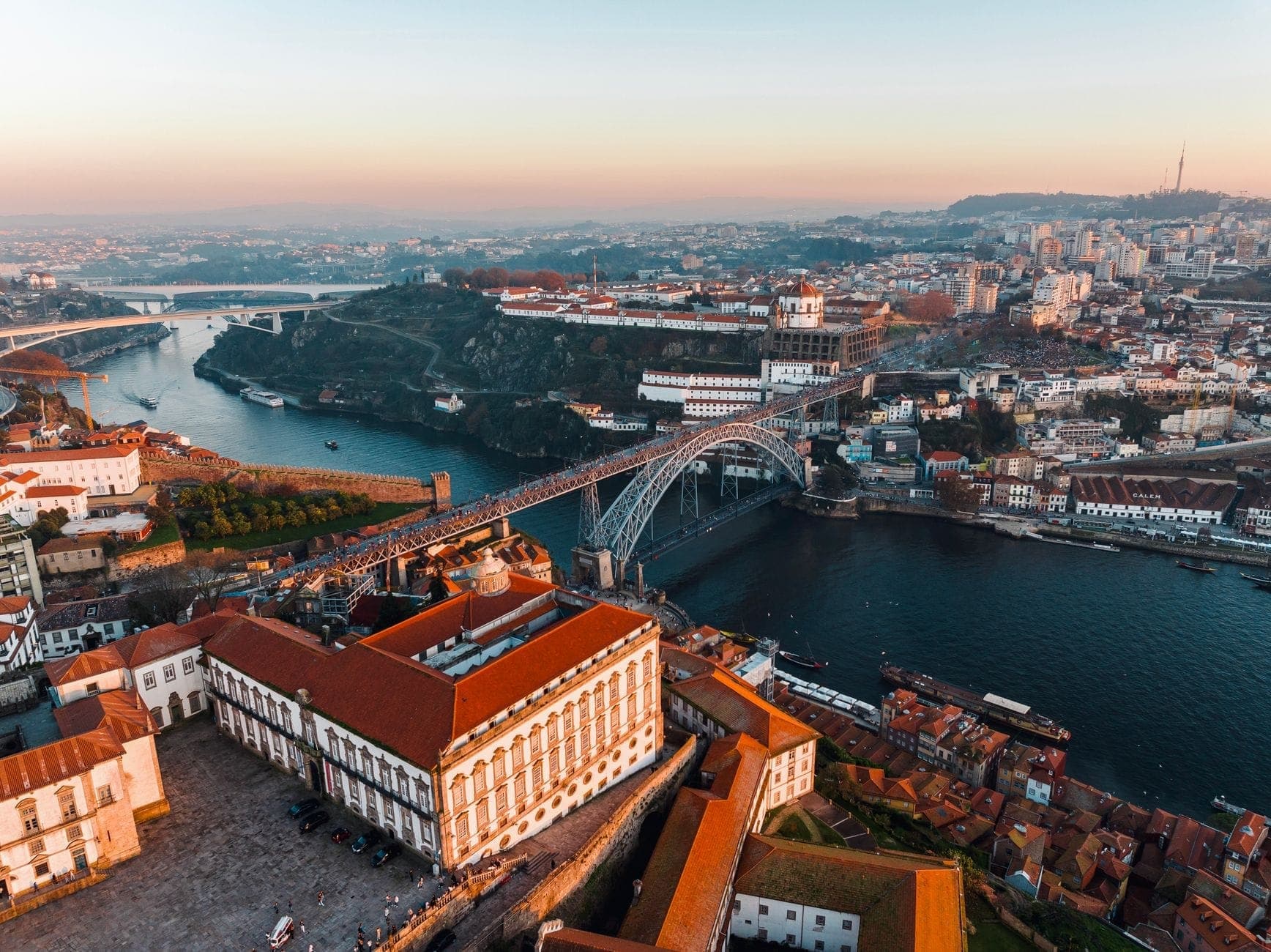 Vista aérea do Porto mostrando a Ponte Dom Luís I, o Rio Douro, edifícios históricos e o Mosteiro da Serra do Pilar ao nascer do sol.