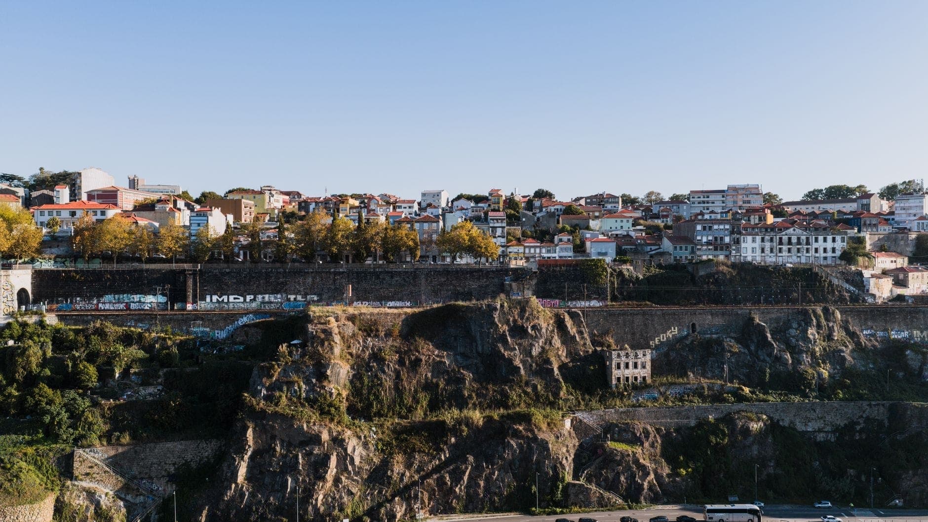 Vista ampla sobre o Porto mostrando penhascos rochosos íngremes abaixo de fileiras de edifícios, destacando o dramático cenário da cidade na encosta.