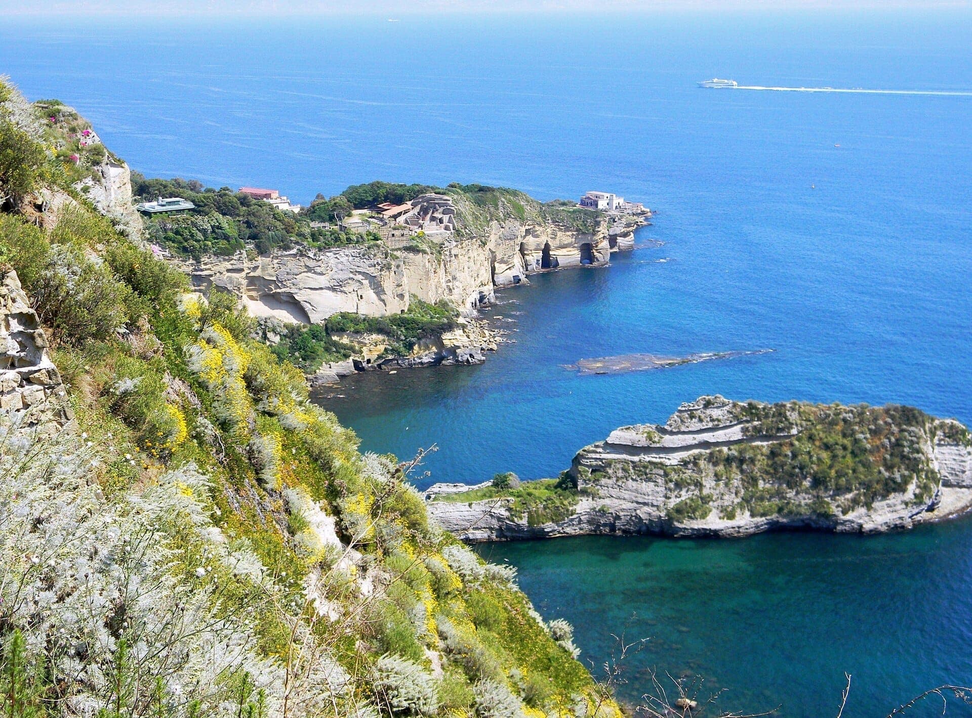 Vista dramática do alto das falésias do Parco Virgiliano sobre a Baía de Nápoles, com mar azul intenso, vegetação exuberante e formações rochosas imponentes lá embaixo.