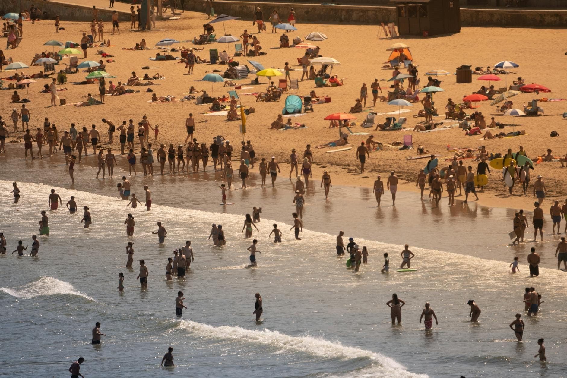Praia de Carcavelos movimentada com banhistas, pessoas tomando sol sob guarda-sóis coloridos e ondas chegando à areia dourada.
