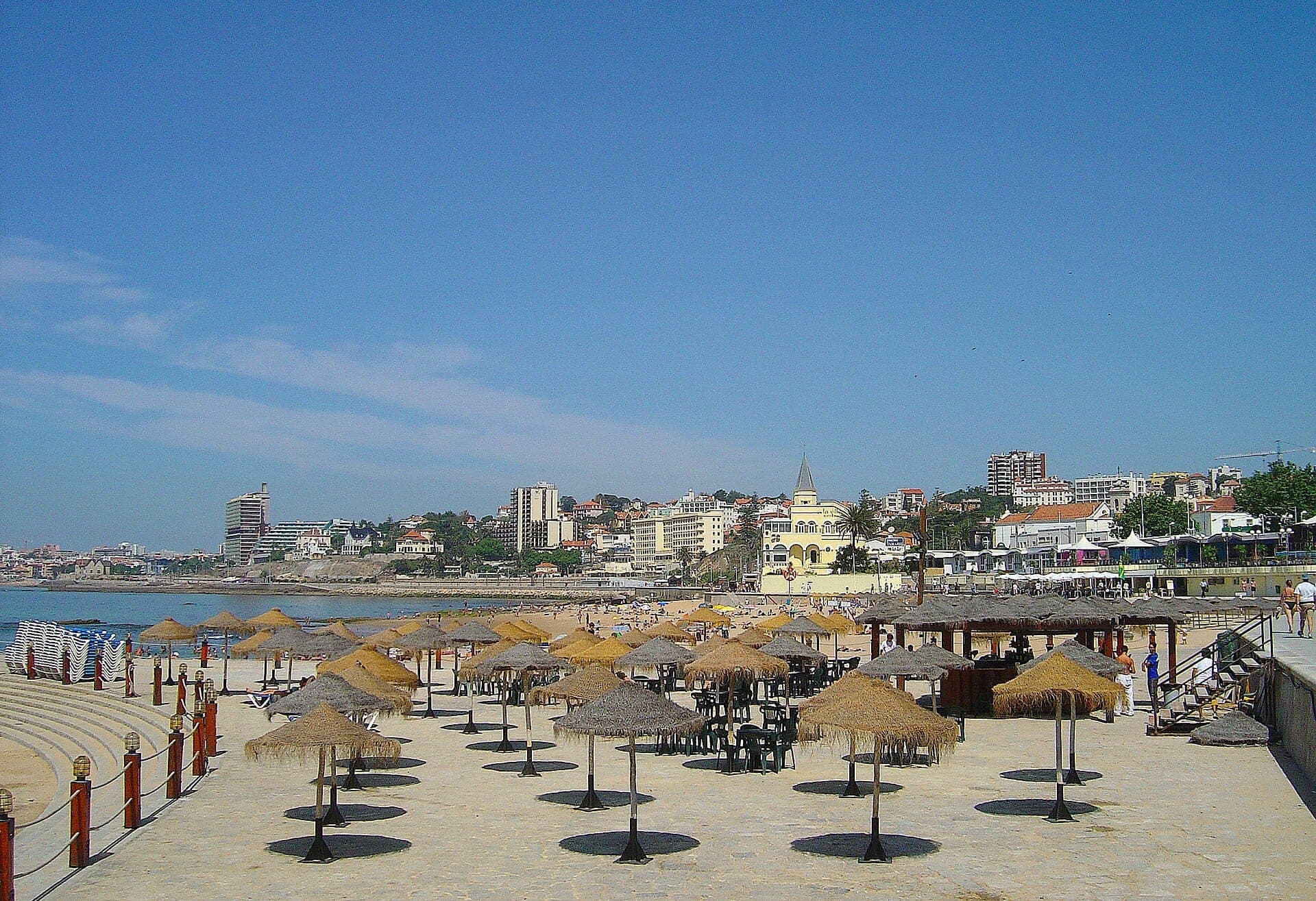 Praia do Tamariz em Estoril com fileiras de chapéus de sol de palha, areia dourada e edifícios da cidade sob um céu azul limpo.