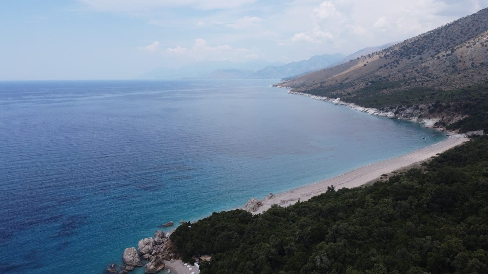 Vue aérienne d'une plage de sable s'étendant sur une étroite bande de terre, entourée d'une mer d'un bleu limpide et d'un littoral sauvage sous un ciel azuré.