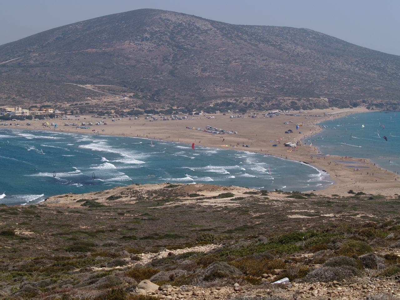 Szeroki widok na plażę Prasonisi na Rodos, gdzie spotykają się dwa piaszczyste brzegi i błękitne morza, z widocznymi windsurferami i plażowiczami.