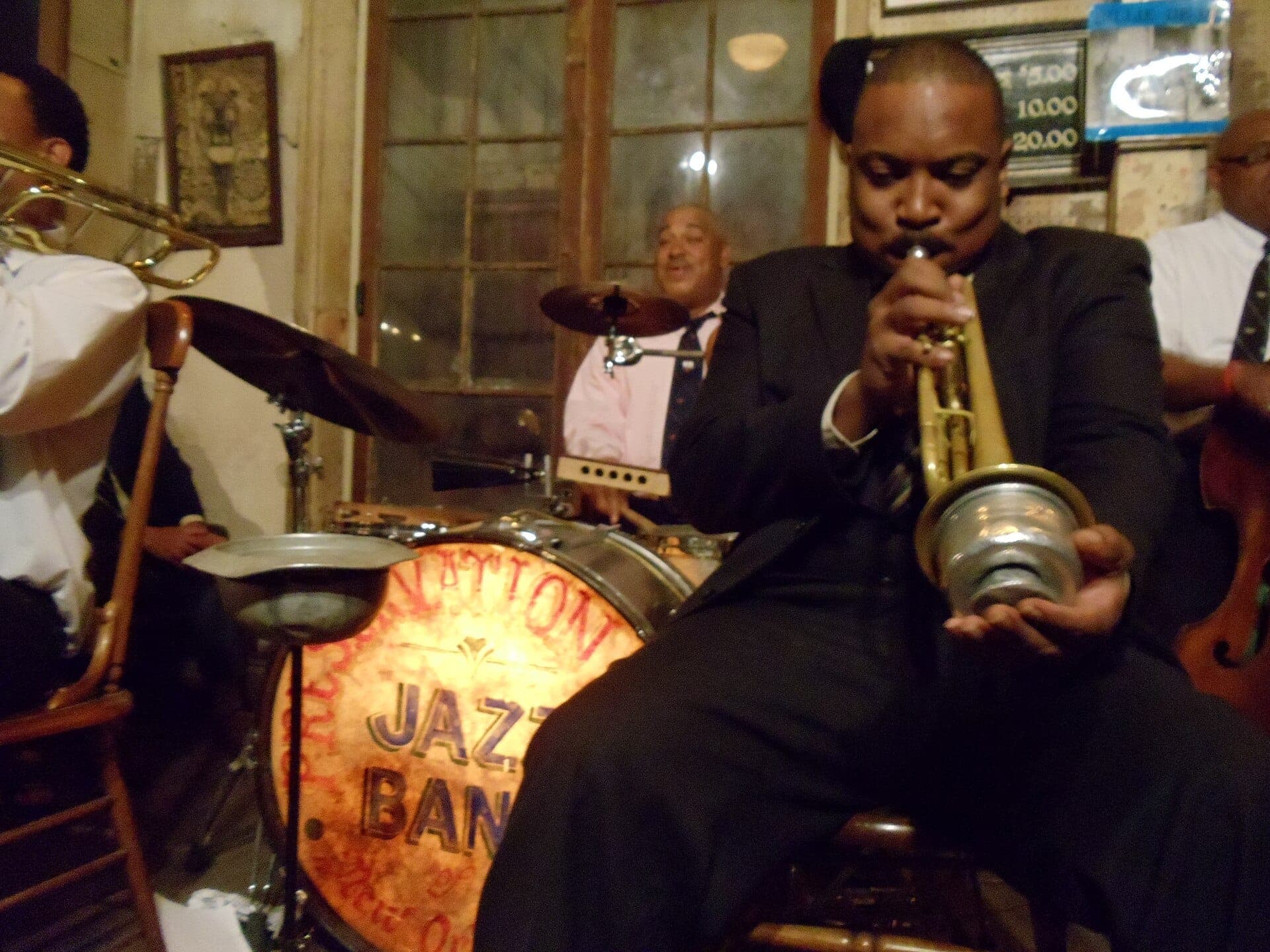 Джазовые музыканты, выступающие вживую в Preservation Hall, с Шэнноном Пауэллом на переднем плане и винтажной барабанной установкой в уютном, слабо освещённом зале.