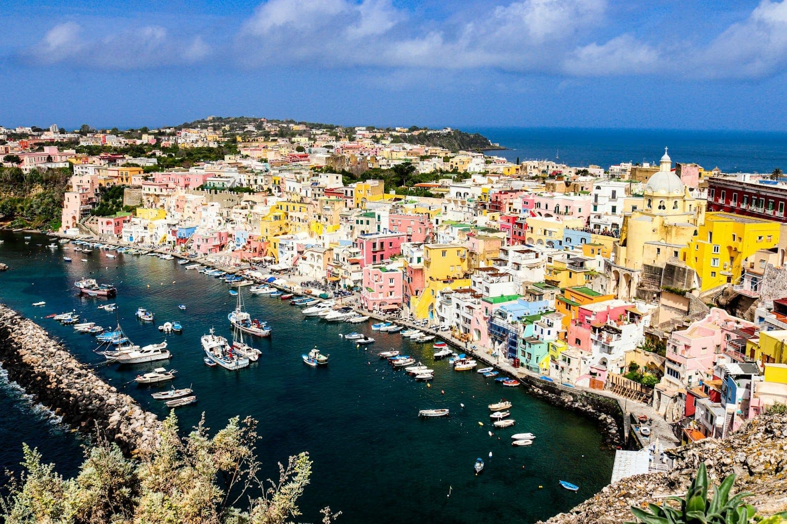Vista geral das coloridas casas em tons pastel do porto da ilha de Procida e barcos de pesca sob a luz do dia, com o mar e o céu azul ao fundo.