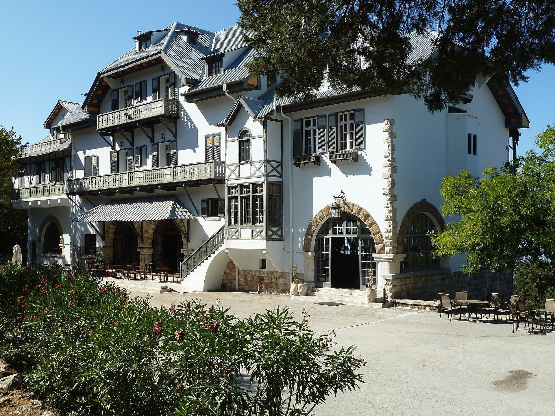 Bâtiment historique de l'époque italienne à Profitis Ilias, Rhodes, entouré d'une végétation luxuriante et de sièges en plein air sous un ciel bleu dégagé.