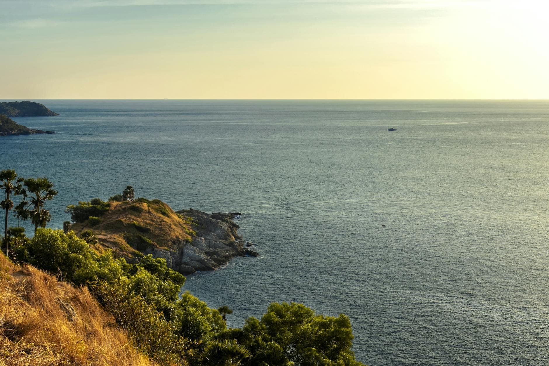Golden hour view of Promthep Cape with rugged headland jutting into calm sea, surrounded by gentle waves, dramatic horizon, and lush foreground foliage.