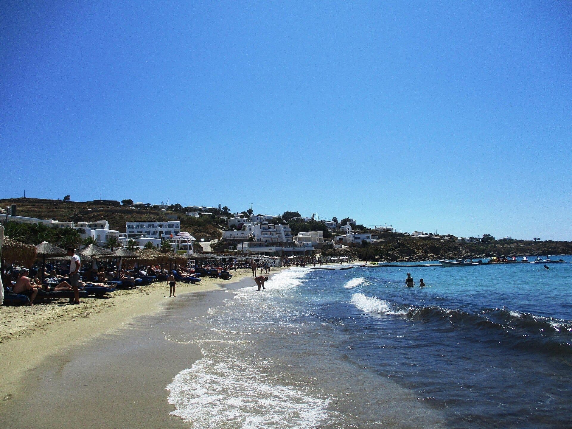 Sonniger Tag am Strand Platis Gialos auf Mykonos, mit Menschen, die sich auf Sonnenliegen entspannen, klarem blauem Meer und weiß getünchten Gebäuden am Hang.