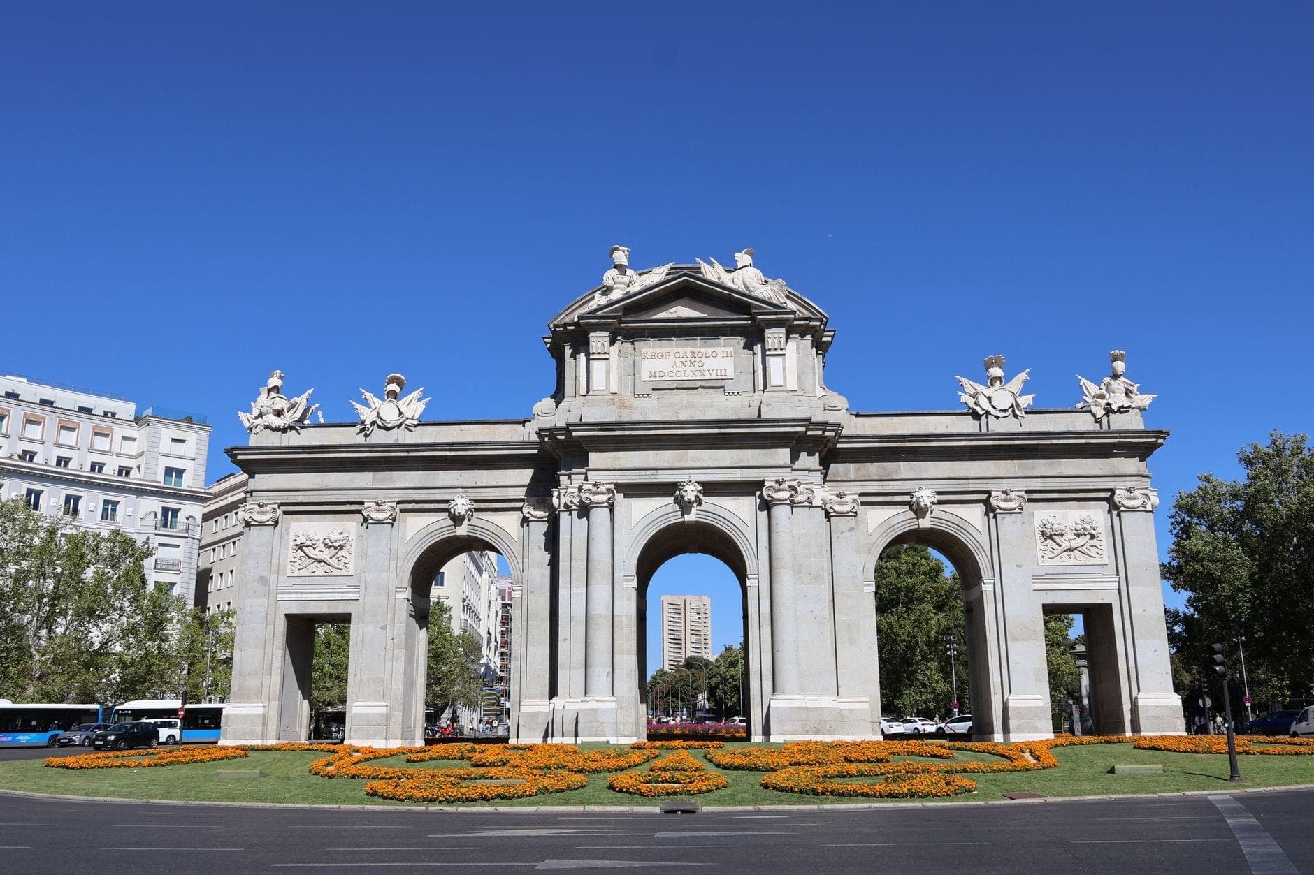 ภาพสดใส ซุ้มประตู Puerta de Alcalá ในมาดริด ท้องฟ้าสีฟ้า ประติมากรรมประดับและแปลงดอกไม้สีส้มอยู่หน้าฉาก ล้อมรอบด้วยตึกกลางเมือง