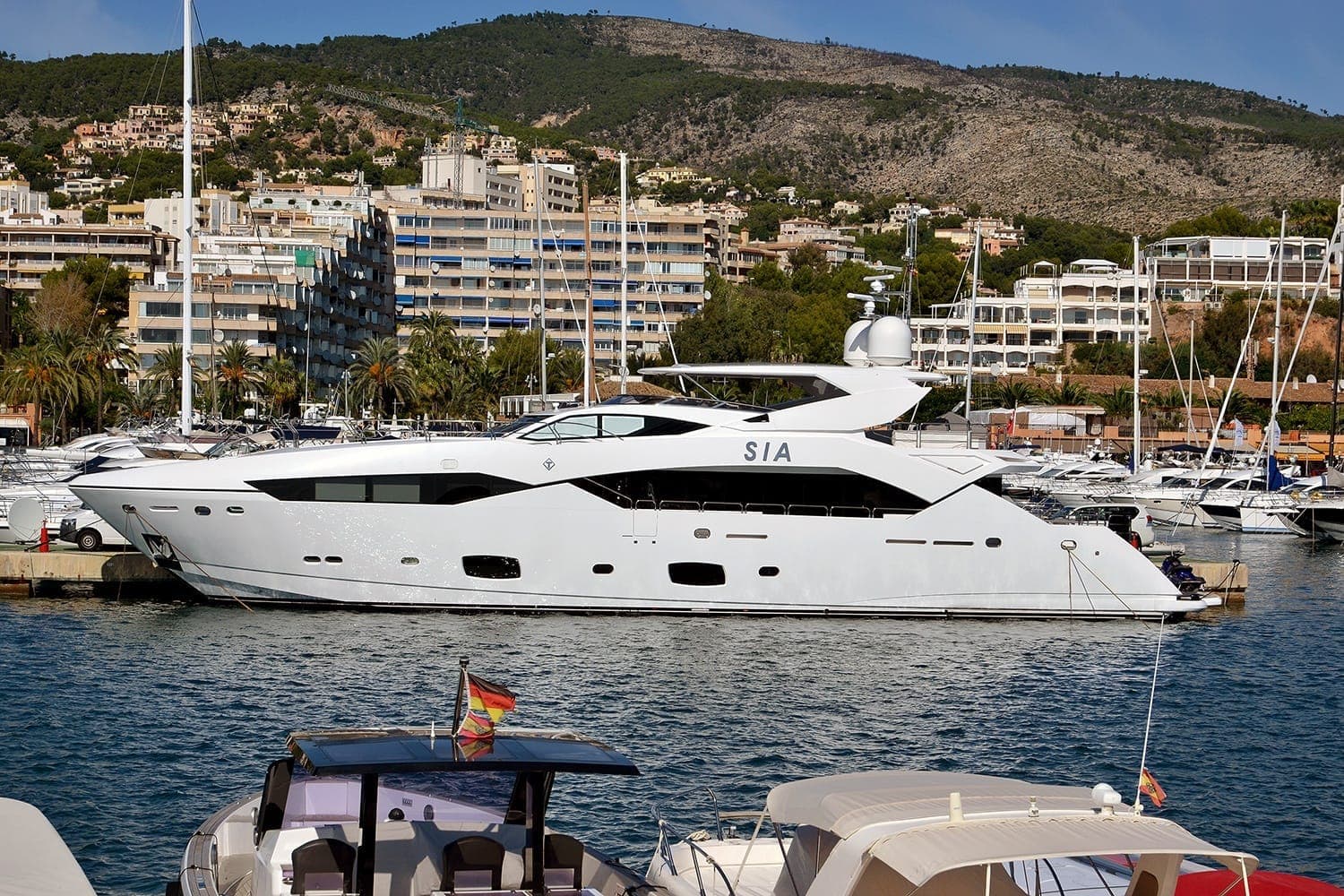 Große weiße Yacht im Hafen von Puerto Portals mit modernen Apartmentgebäuden, grünen Hügeln und Palmen im Hintergrund bei hellem Tageslicht.