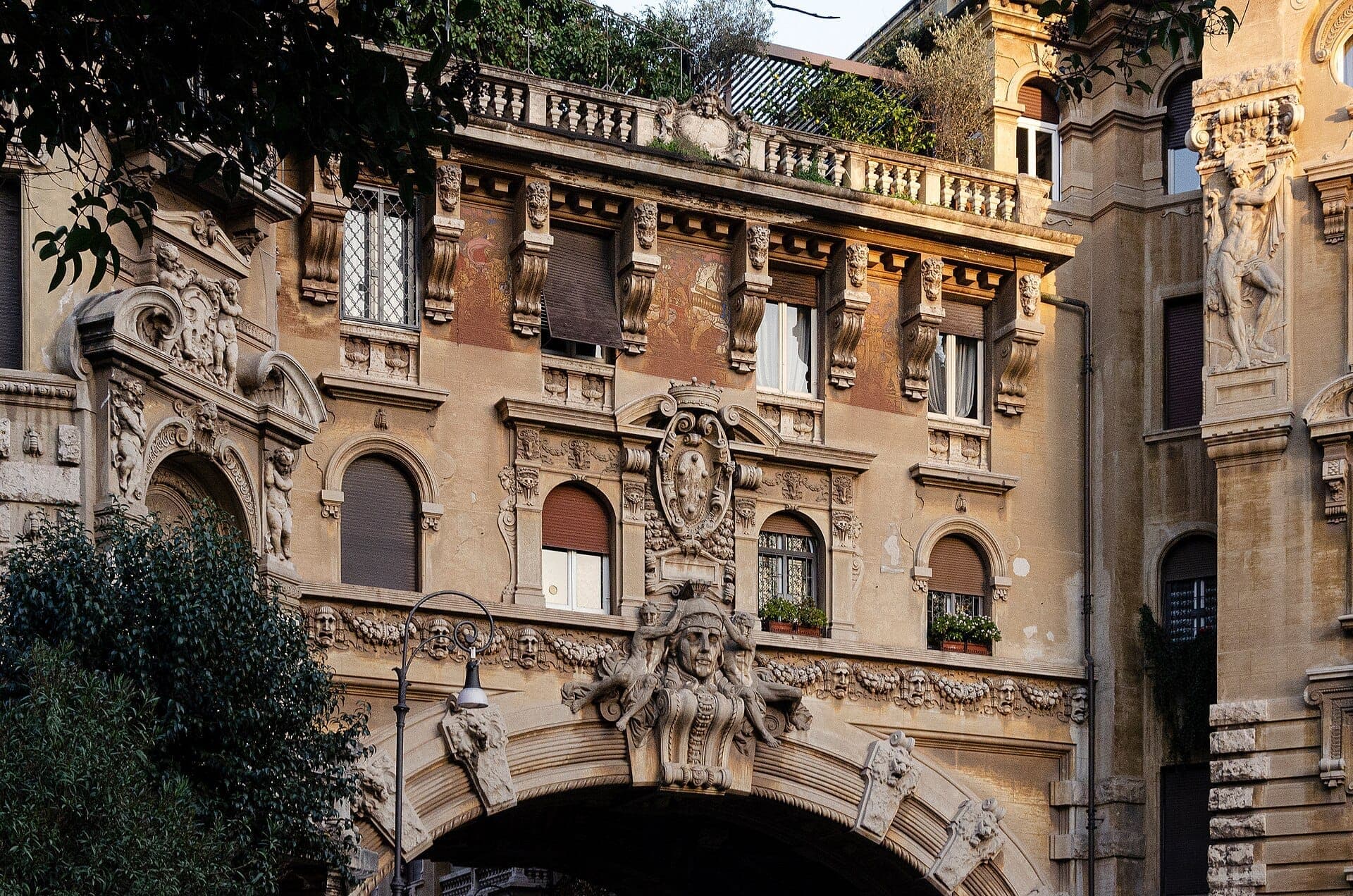 ภาพระยะใกล้ของด้านหน้าอาคารประดับประดาและซุ้มโค้งหินตกแต่งของ Quartiere Coppedè ที่มีหน้าแกะสลักซับซ้อนและต้นไม้บนระเบียง ใต้แสงกลางวันอบอุ่น