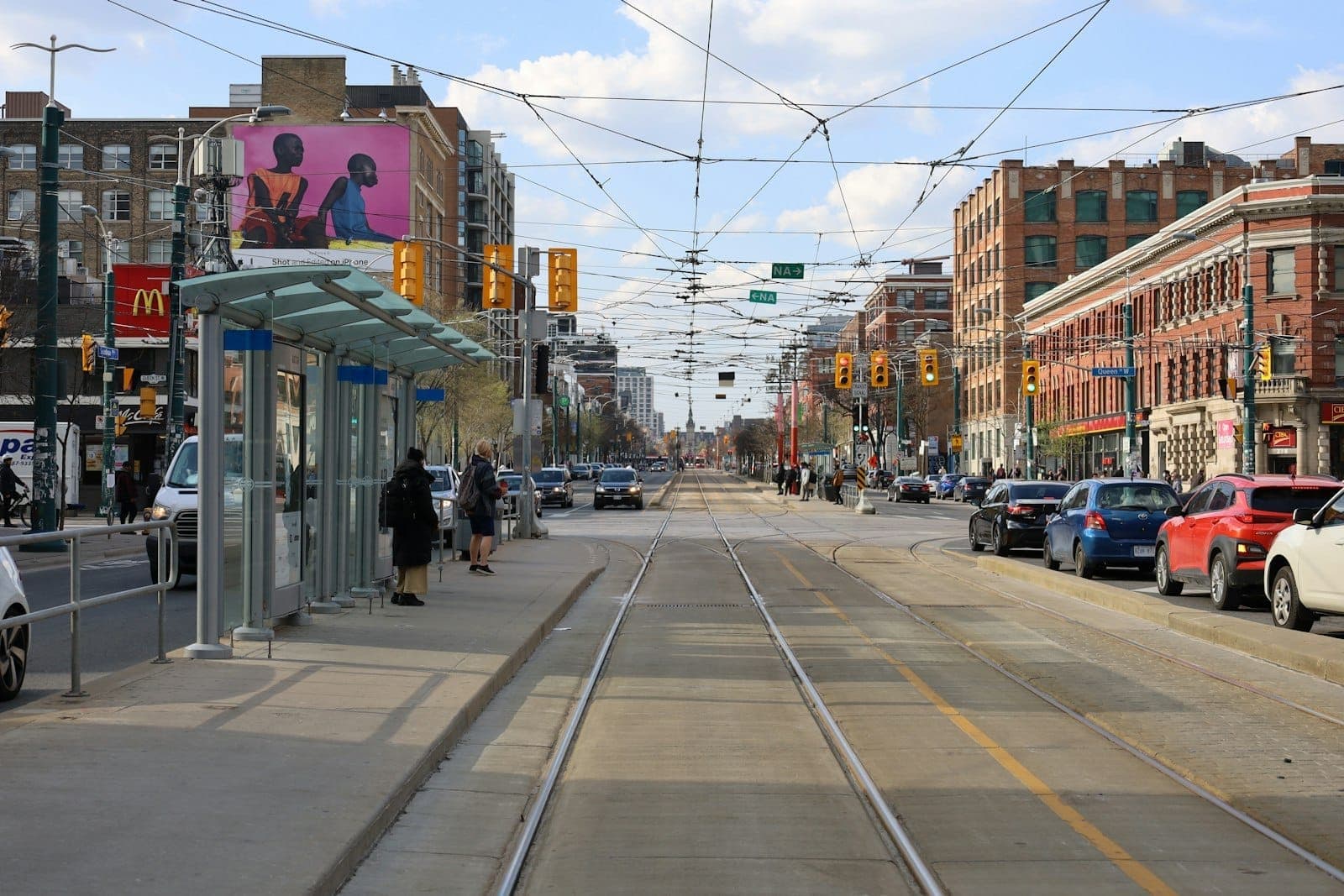Vista diurna di Queen Street West a Toronto con i binari del tram, persone alla fermata, edifici storici in mattoni e un'atmosfera urbana vivace.