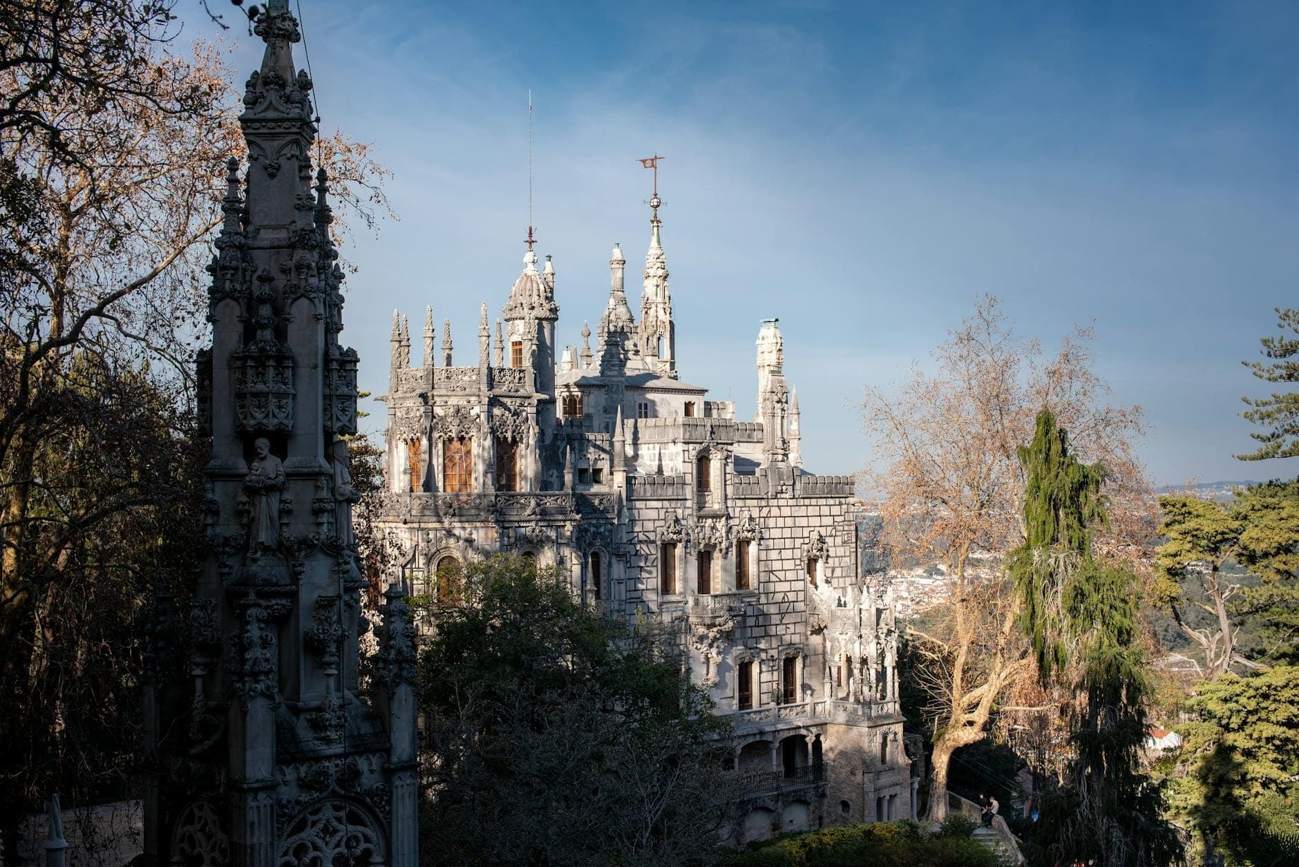Vista ampla do palácio da Quinta da Regaleira com sua ornamentada arquitetura neo-gótica cercada por jardins exuberantes e árvores sob um céu azul em Sintra, Portugal.