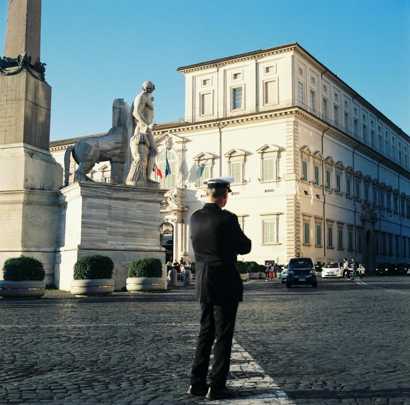 Pałac Kwirynalski w Rzymie — przy obelisku i posągach stoi wartownik w mundurze, na tle bezchmurnego niebieskiego nieba.