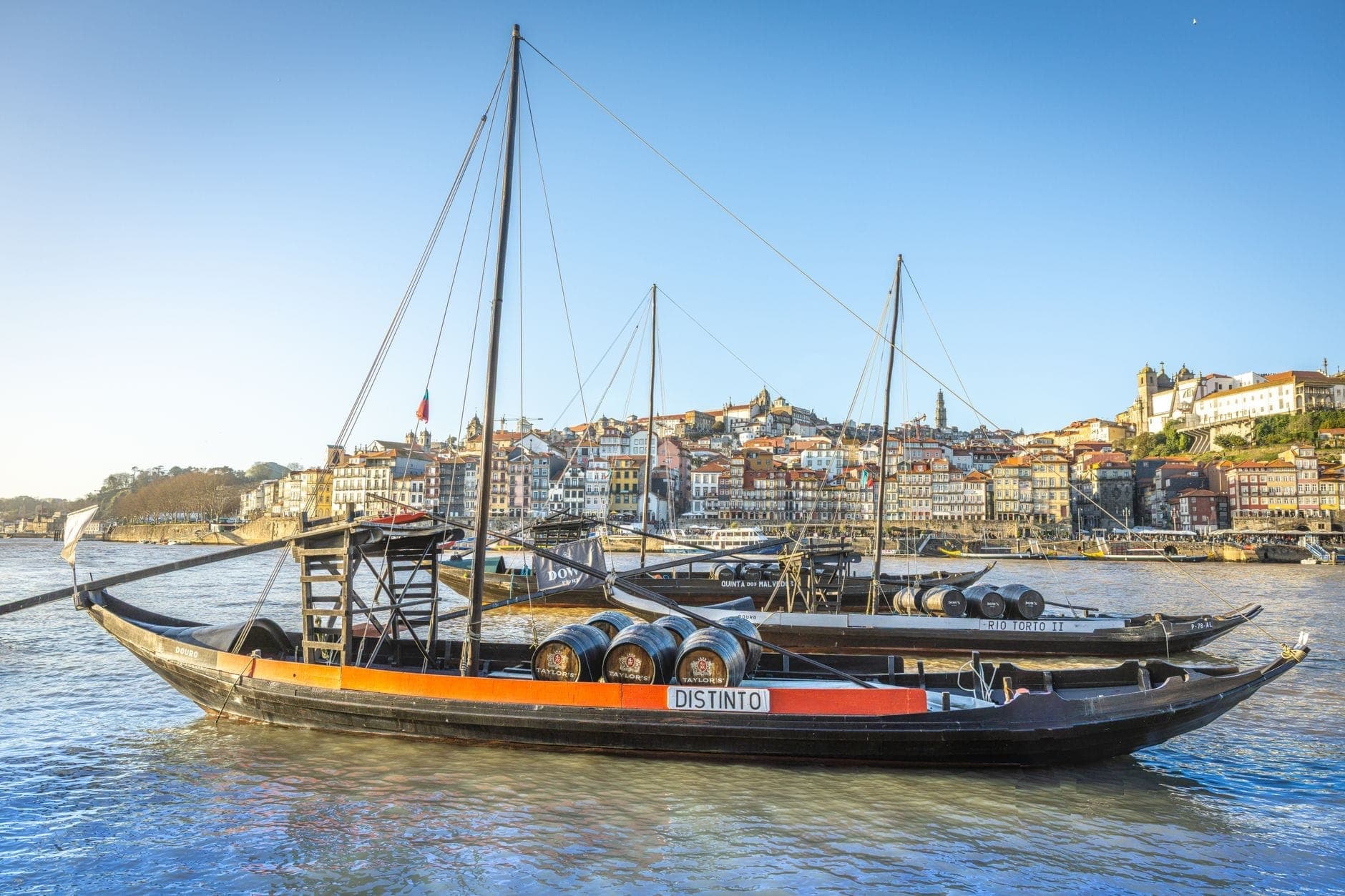Bateaux rabelo traditionnels chargés de tonneaux sur le Douro, avec la ville de Porto et ses façades colorées en arrière-plan sous un ciel bleu.