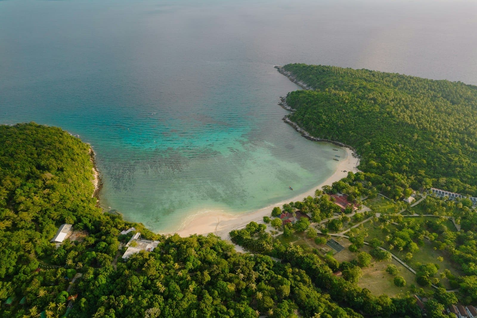 Aerial view of Koh Racha Yai’s white sandy beach curving between lush green hills and turquoise water, with a few buildings and boats visible.