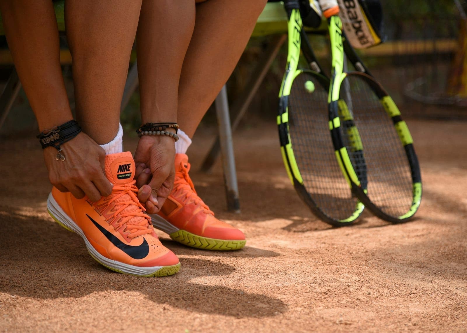 Nahaufnahme einer Person, die orangefarbene Tennisschuhe neben zwei gelben Tennisschlägern auf einem Sandplatz im Rafa Nadal Museum Xperience schnürt.