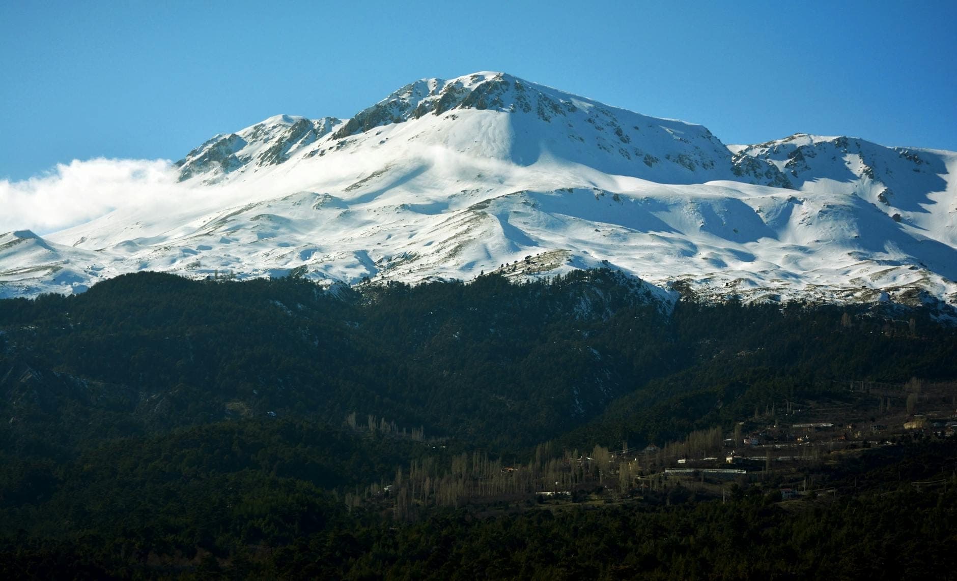 Schneebedeckte Taurus-Berge unter einem klaren blauen Himmel, bewaldete Unterhänge mit einigen verstreuten Gebäuden.