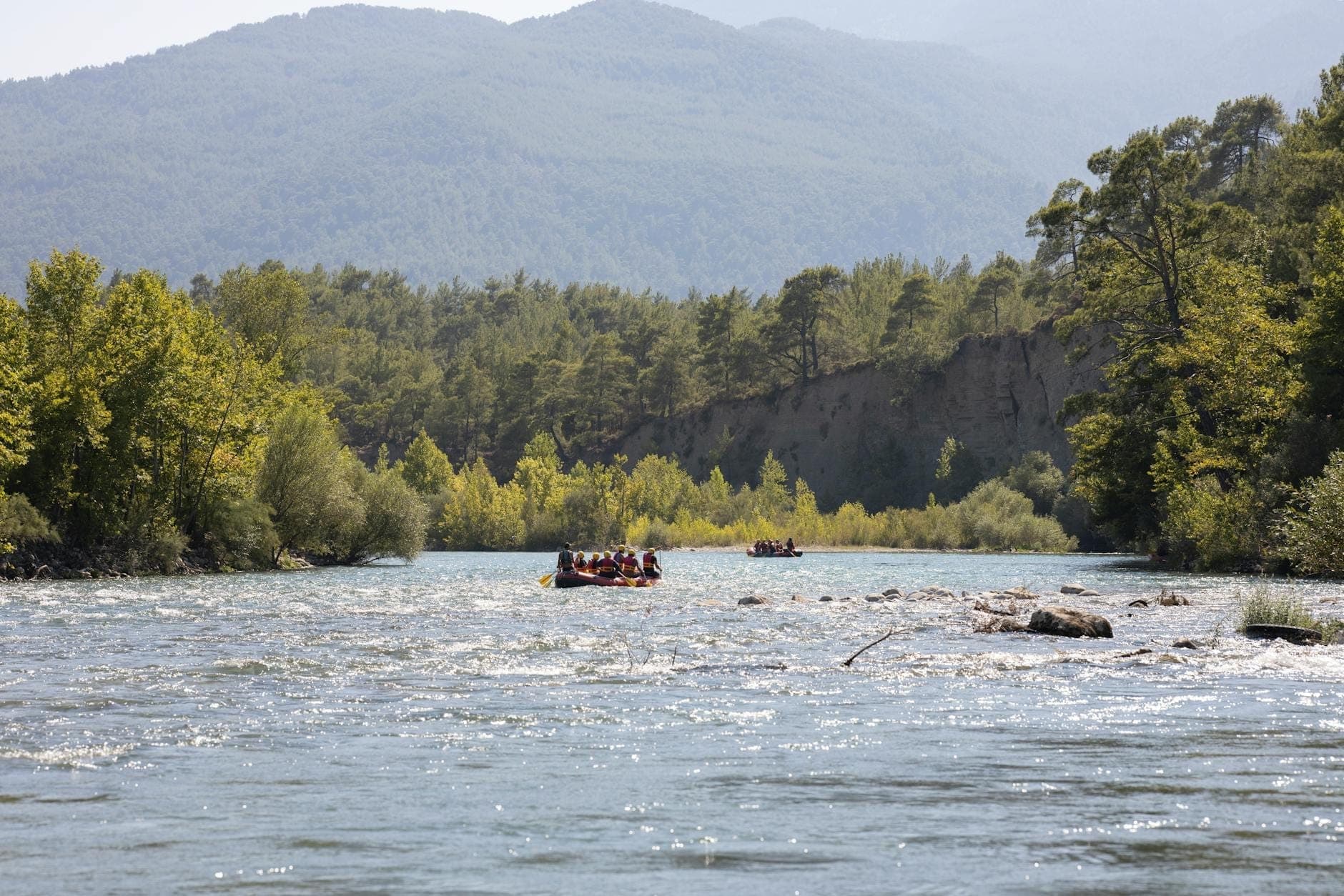 Weitwinkelansicht von Flößen, die einen malerischen Fluss hinuntertreiben, umgeben von bewaldeten Ufern und einer fernen Berglandschaft.