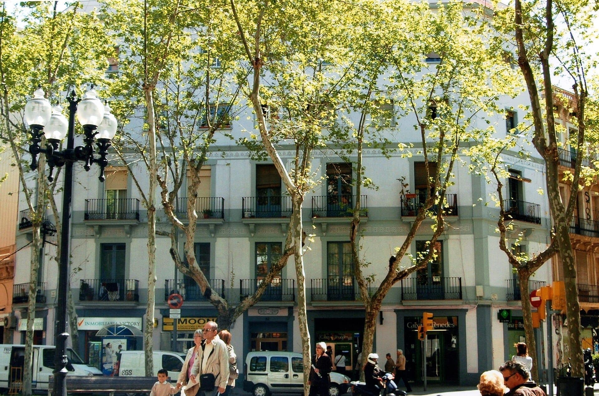 ทางเดินเรียงต้นไม้บน Rambla del Poblenou พร้อมอาคารเตี้ยสไตล์คลาสสิก กิ่งไม้ใบดกให้ร่มเงาแสงแดดเป็นจุดๆ และชาวบ้านเดินเล่นอยู่เบื้องล่าง