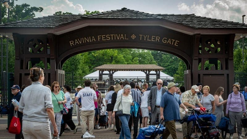 Los visitantes entran por la puerta de madera Ravinia Festival Tyler Gate, rodeada de árboles y nubes, con multitudes que llevan sillas de jardín y suministros para picnic.