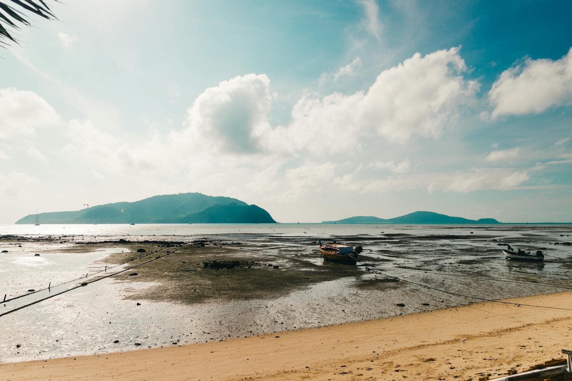 Low tide at Rawai Beach with traditional long-tail fishing boats anchored on the sandy shoreline, distant islands, and a bright blue sky with scattered clouds.