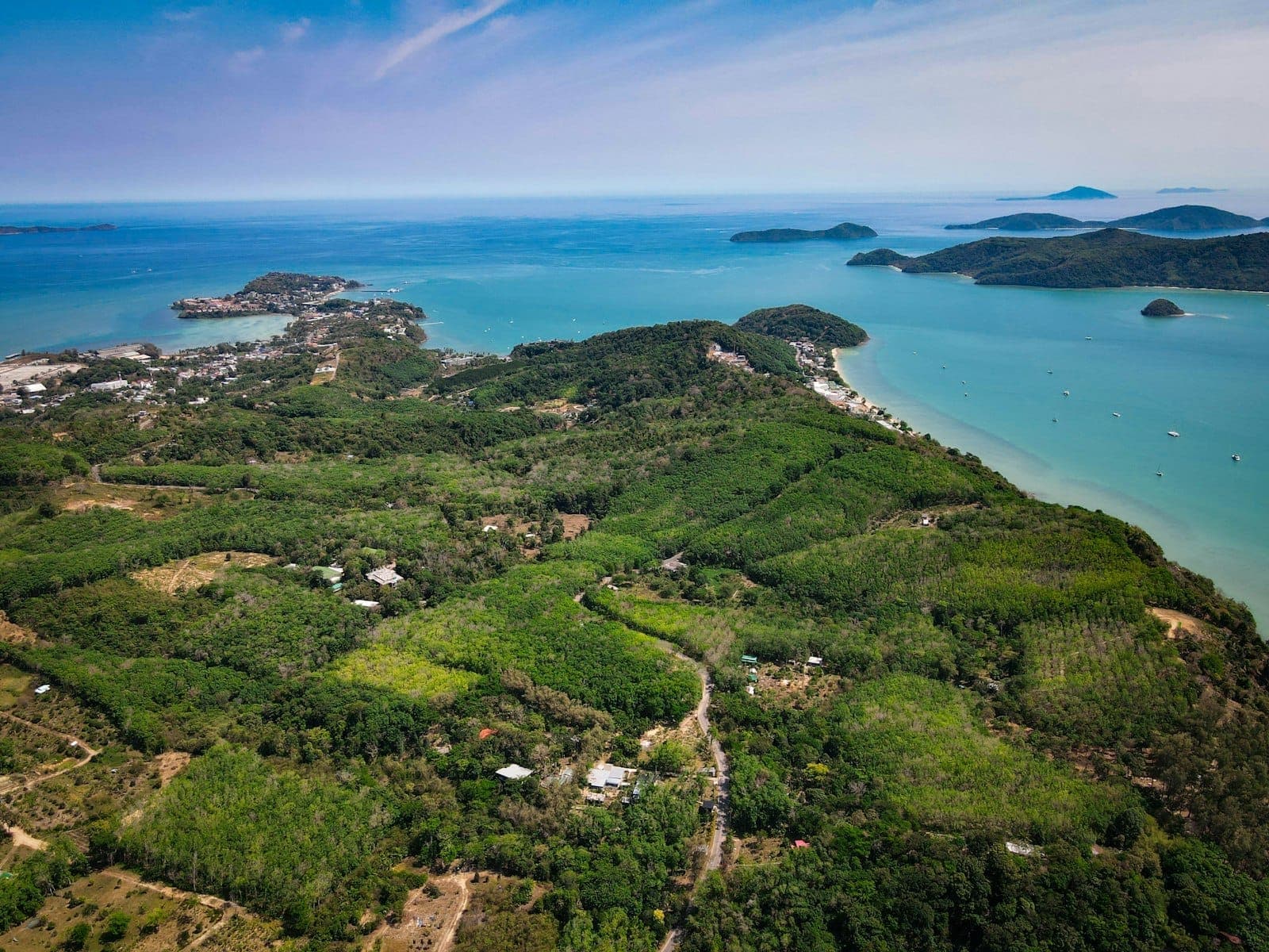 A stunning aerial view of Rawai and Chalong’s coastline with lush green hills, turquoise sea, small islands, and scattered boats under a bright blue sky.