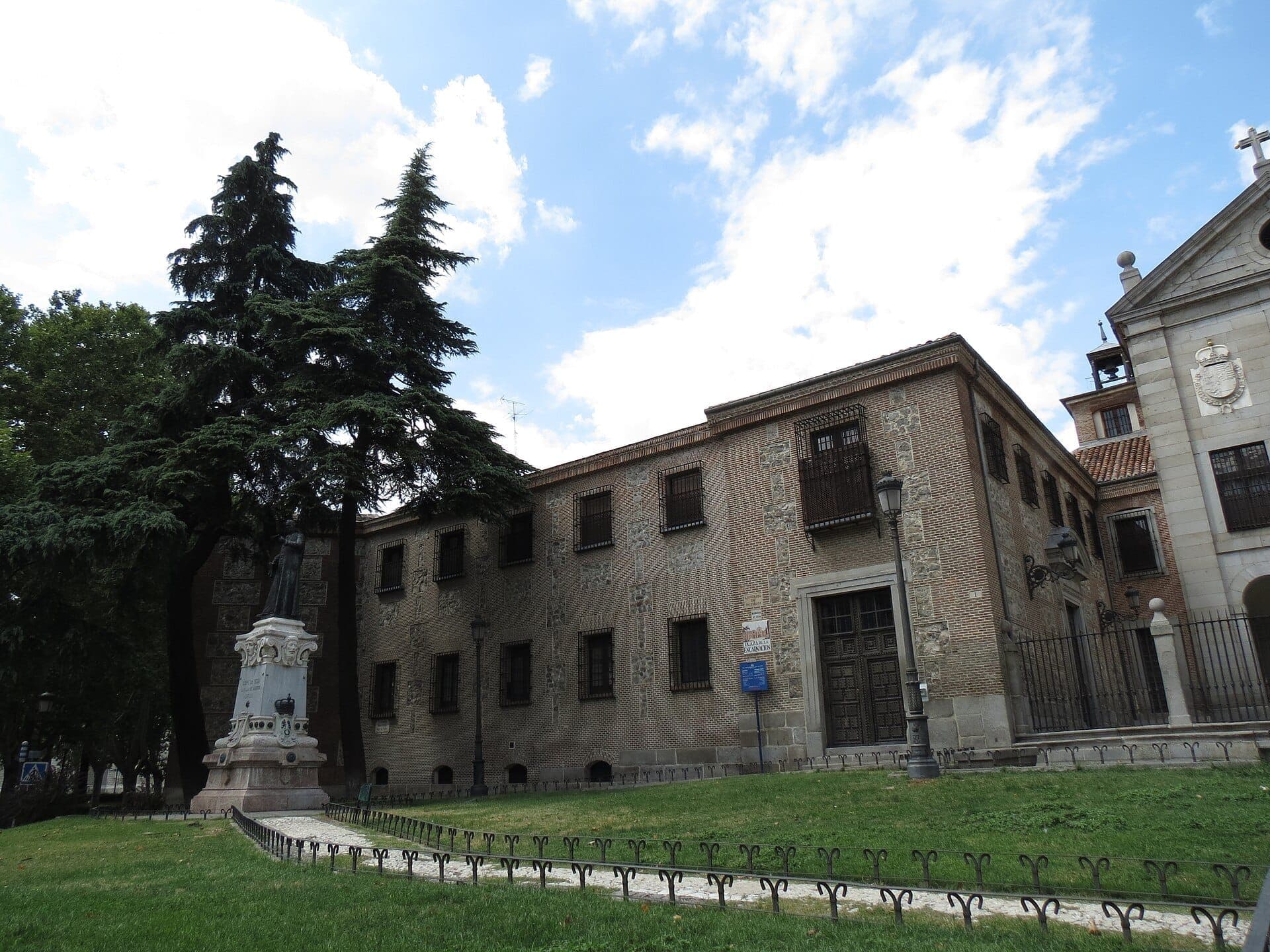 Кирпичный фасад Real Monasterio de la Encarnación в Мадриде со статуей и пышным зелёным газоном под ярко-голубым небом.