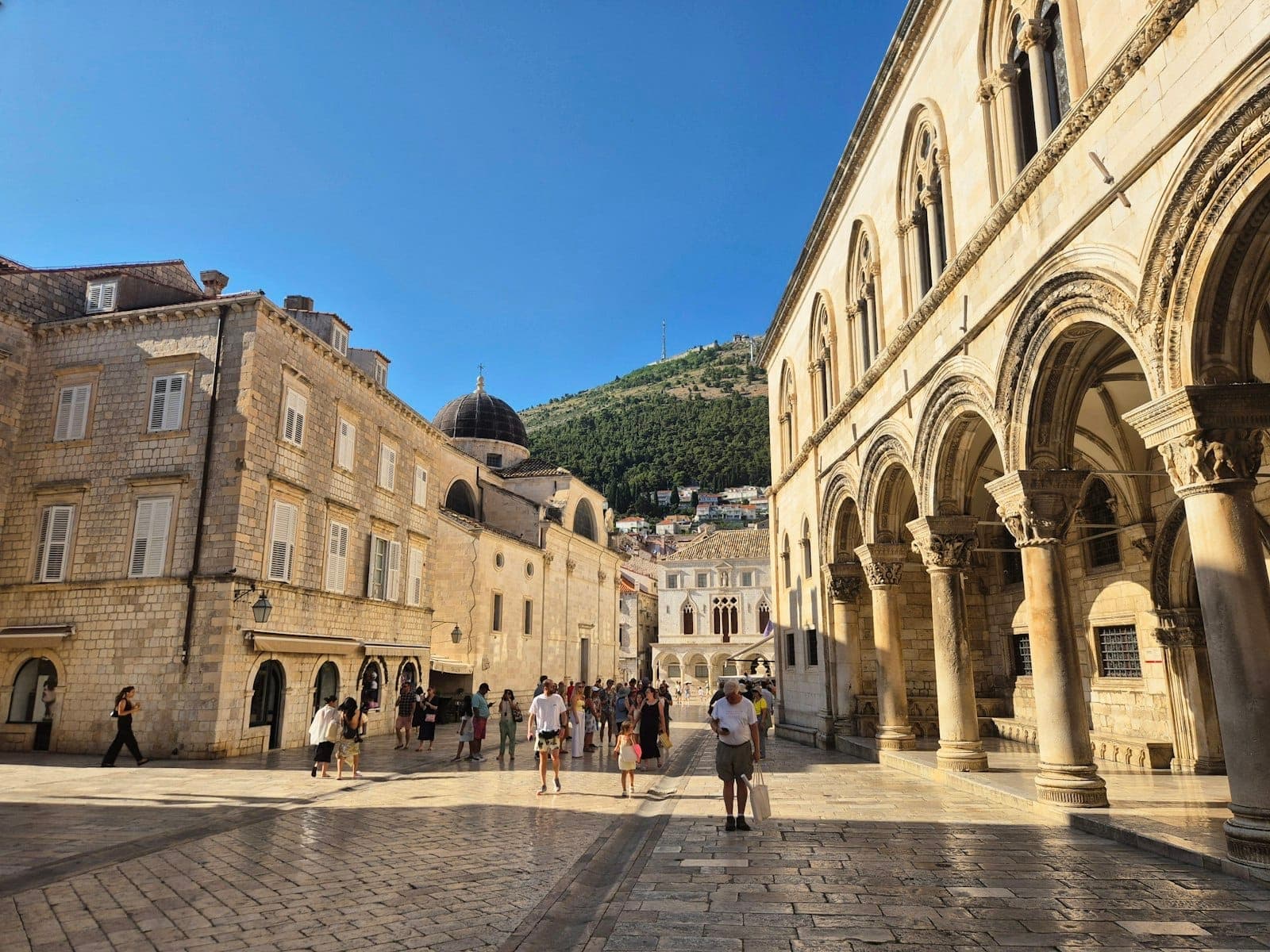 Weitläufige Ansicht des Rektorenpalasts in Dubrovnik mit eleganten Bögen, historischen Nachbargebäuden, Besuchern auf dem sonnenbeschienenen Altstadtplatz und Hügeln im Hintergrund.