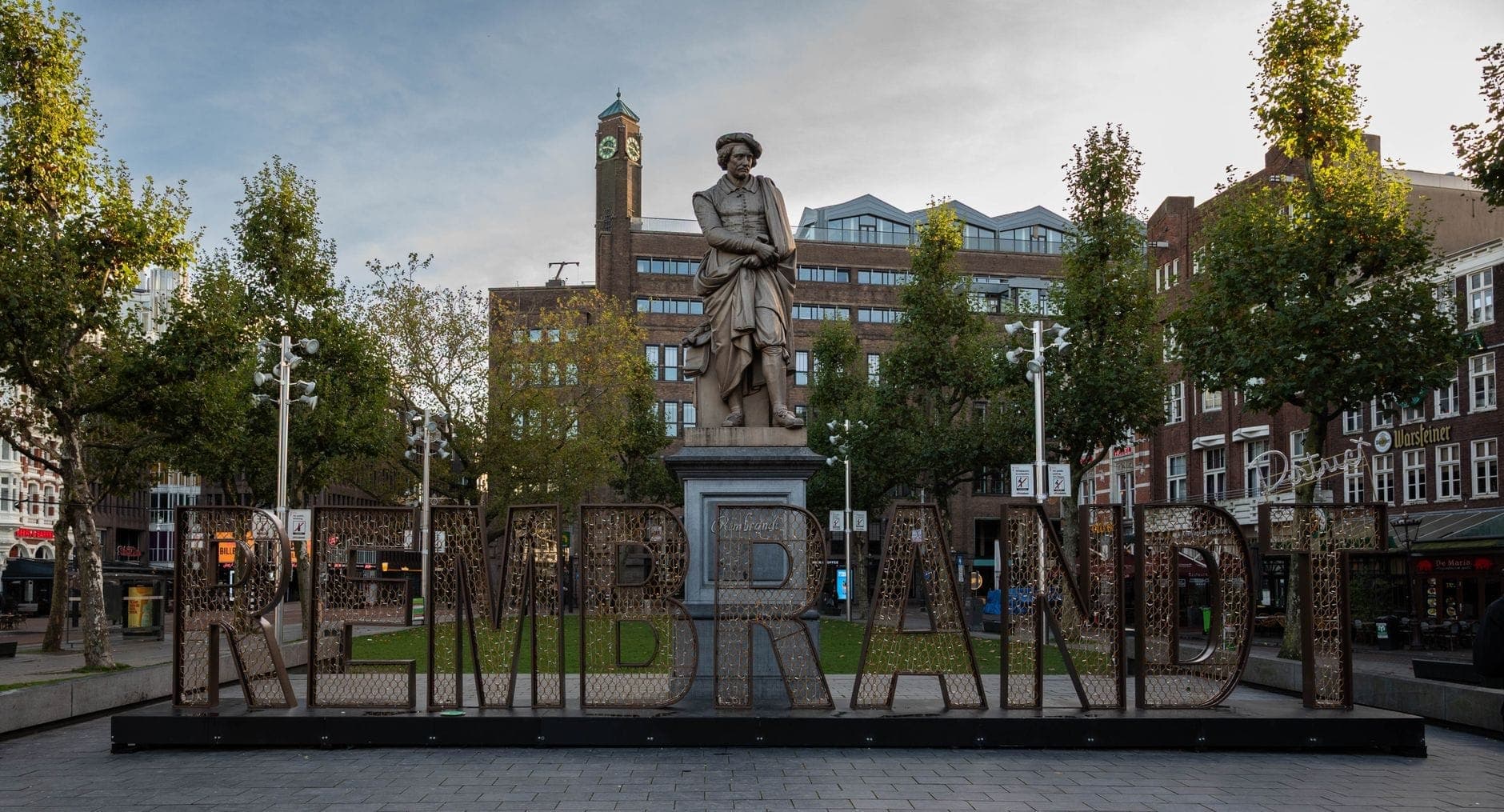 Die Rembrandt-Statue steht im Zentrum des Rembrandtplein in Amsterdam, umgeben von Bäumen, historischen Gebäuden und offenem Platz unter einem teils bewölkten Himmel.