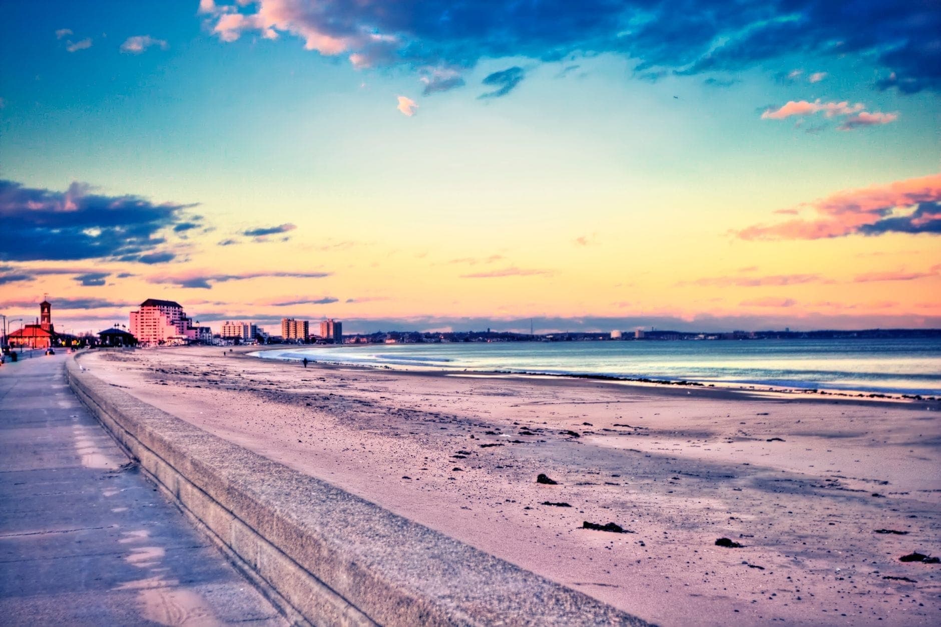 Vue panoramique sur une longue plage de sable longeant une large promenade, avec les vagues de l'océan et les bâtiments de la région de Boston au loin sous un ciel coloré au coucher du soleil.
