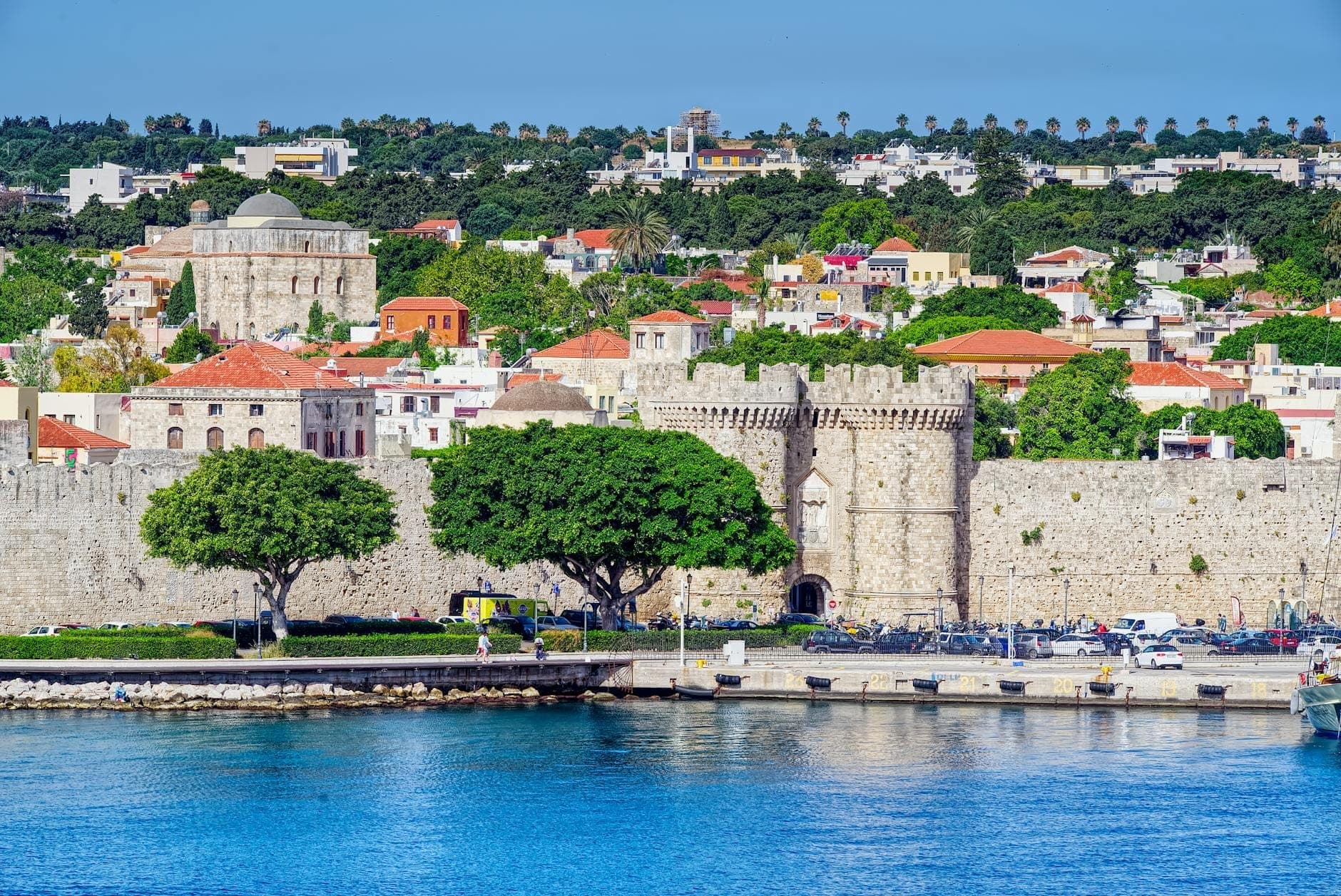 Vue animée de la ville de Rhodes avec ses remparts médiévaux en pierre, ses bâtiments historiques, ses arbres luxuriants et une mer d'un bleu éclatant au premier plan sous un ciel dégagé.