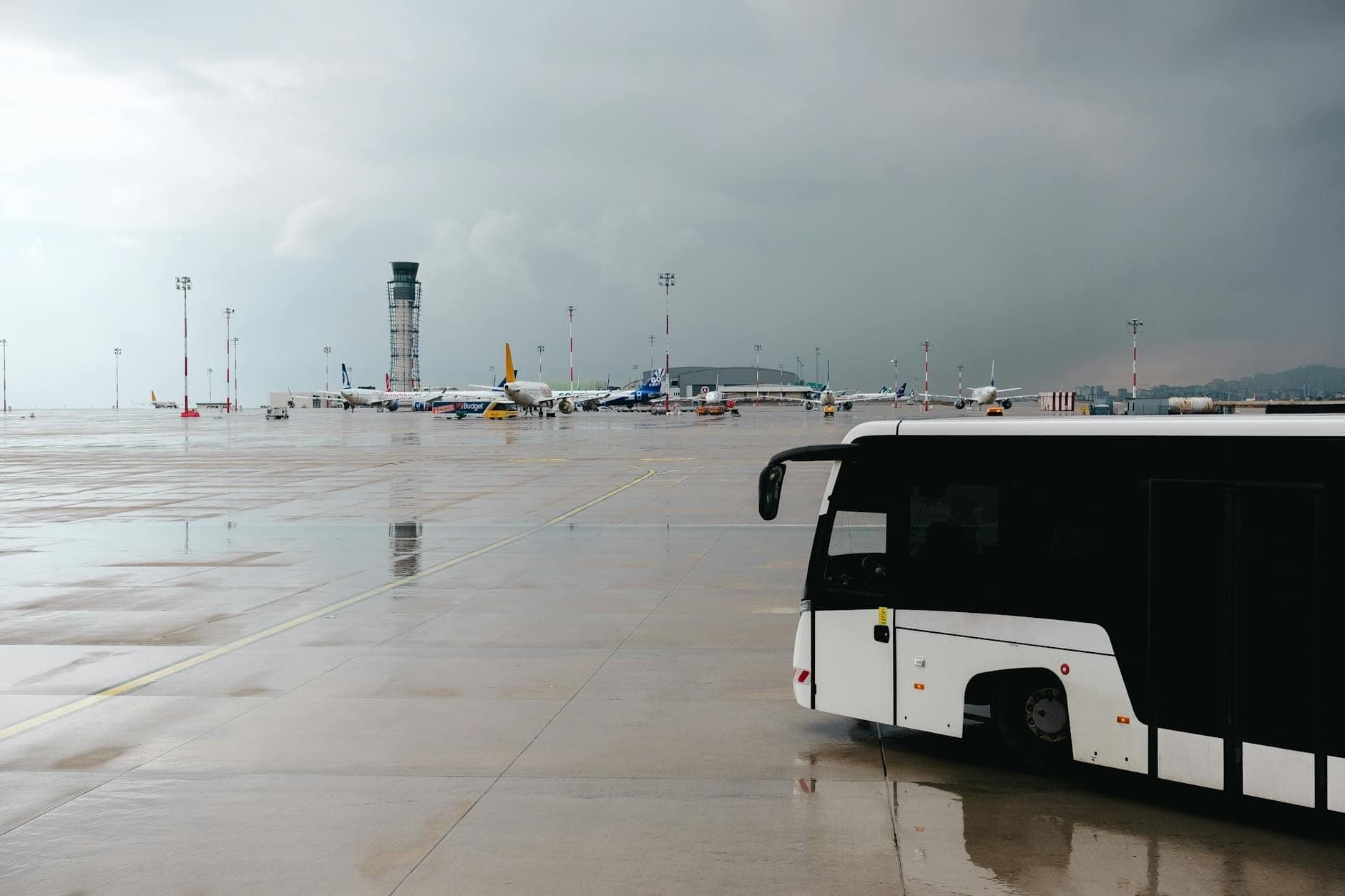 Un bus blanc d'aéroport sur le tarmac avec des avions et une tour de contrôle visibles en arrière-plan sous un ciel nuageux.