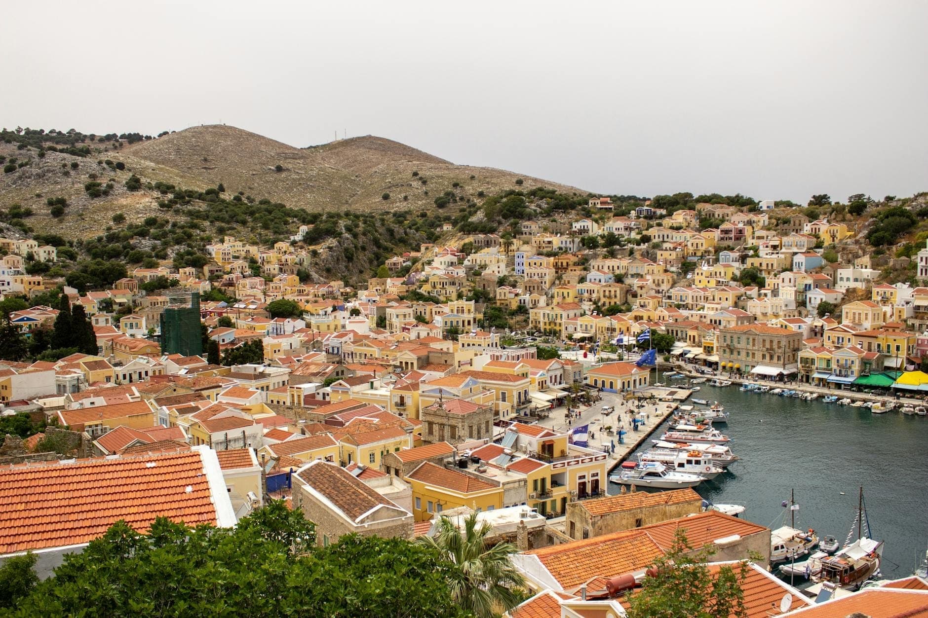 Widok z lotu ptaka na kolorowy neoklasyczny port i domy na wzgórzu Symi Island, łodzie cumujące przy nabrzeżu, pod zachmurzonym niebem.