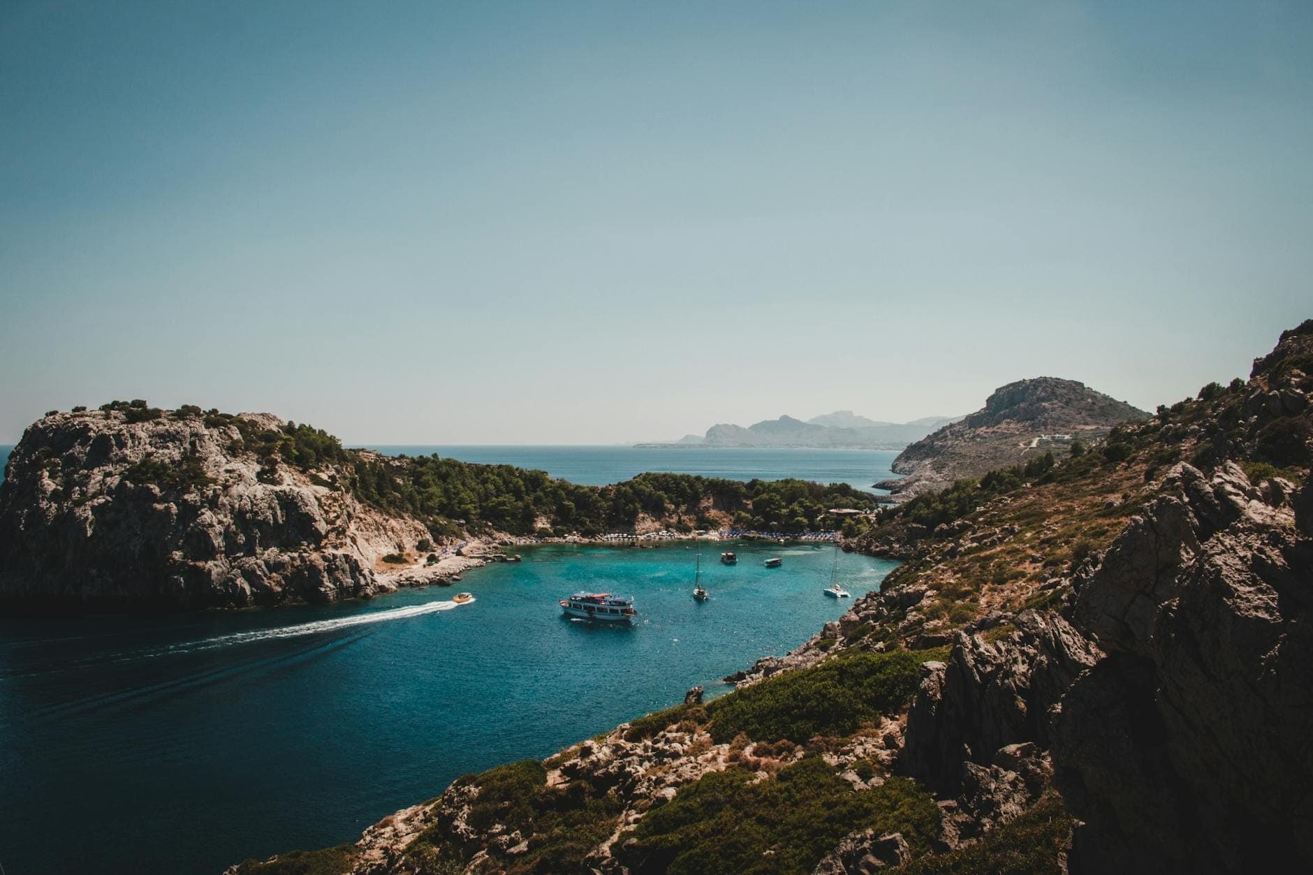 Une crique côtière pittoresque à Rhodes encadrée de collines rocheuses, avec une eau bleue cristalline, des bateaux au mouillage et des montagnes au loin sous un ciel lumineux.