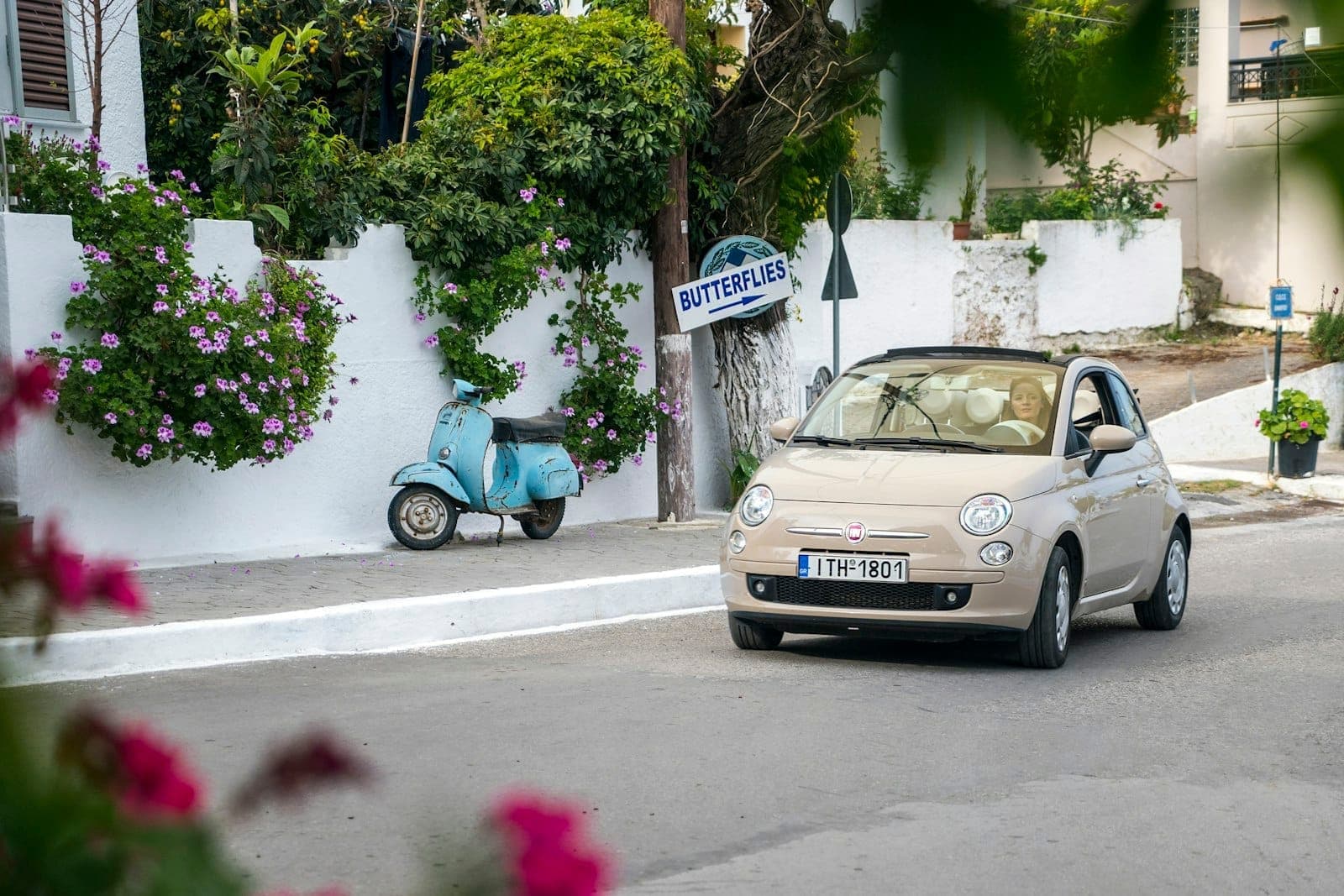 Une Fiat 500 beige passe devant un mur blanchi à la chaux avec des fleurs roses, un scooter bleu vintage et un panneau indiquant 'Butterflies' à Rhodes.