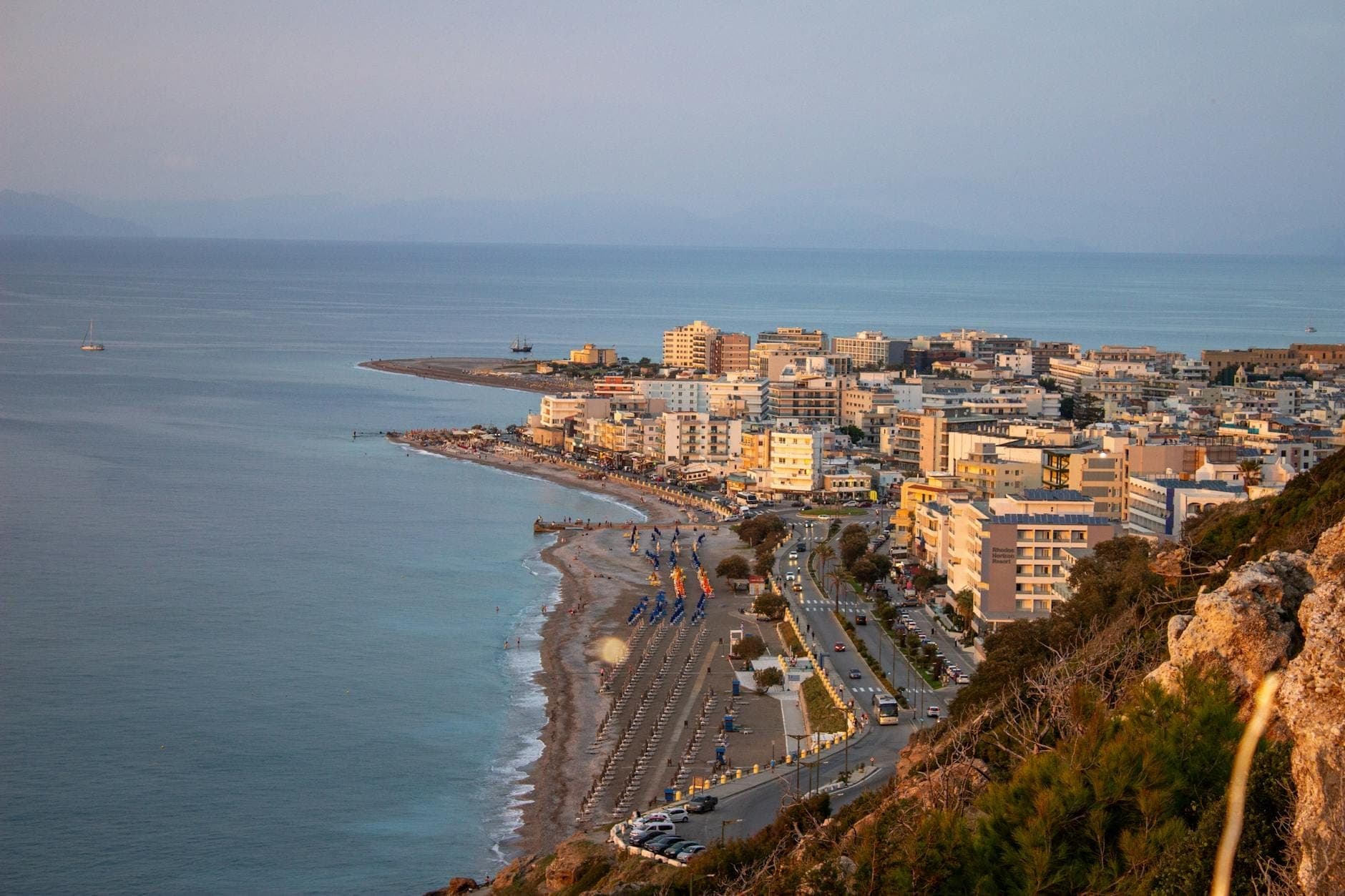 Vue en hauteur du littoral et de la ville de Rhodes, avec des bâtiments s'étendant le long du rivage et la mer au coucher du soleil, vue depuis un point de vue élevé.