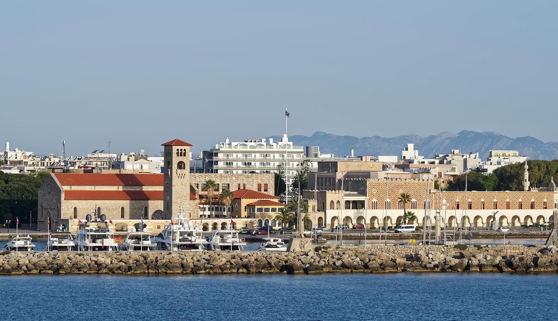 Vue du front de mer du port de Mandraki avec des bâtiments gouvernementaux de style italianisant et un beffroi prominent, des yachts amarrés le long de la promenade et des collines en arrière-plan.