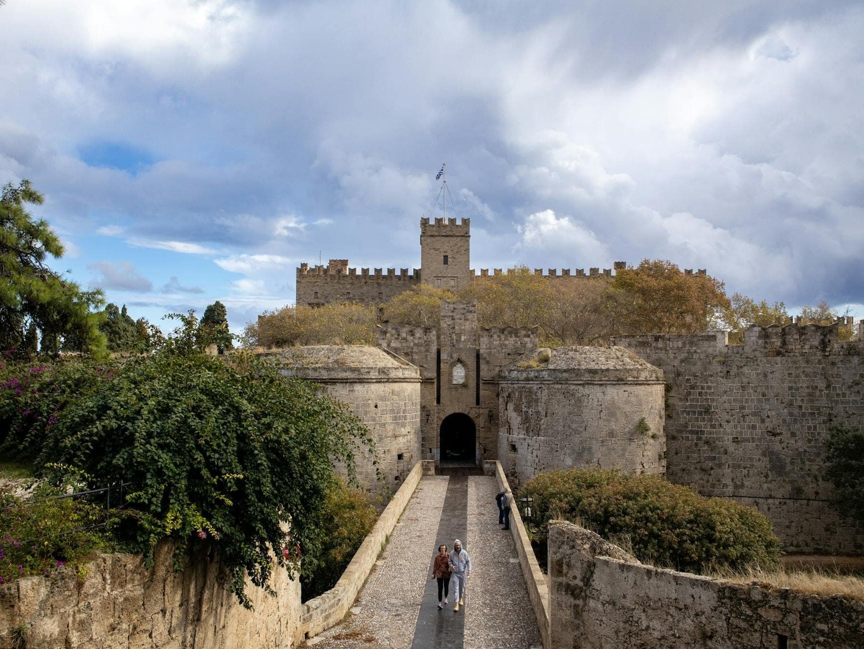 Remparts et entrée d'un château médiéval en pierre, avec un chemin entouré d'une végétation luxuriante sous un ciel dramatique à Rhodes, en Grèce, évoquant une histoire enfouie.