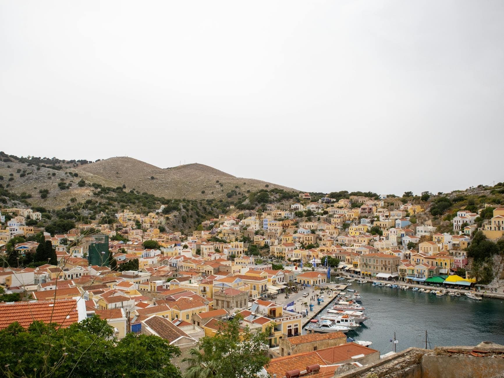 Vue panoramique d'une ville portuaire avec des maisons colorées, des bateaux amarrés le long du front de mer et des collines arides s'élevant en arrière-plan.