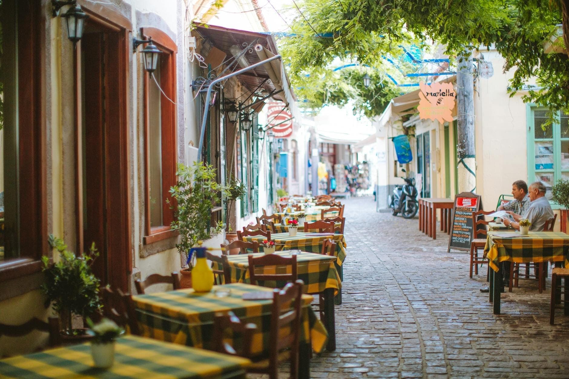 Urocza ulica dla pieszych na Rodos obsadzona stolikami kawiarni na świeżym powietrzu pod zielonymi drzewami, z jedzącymi ludźmi i kolorowymi budynkami.