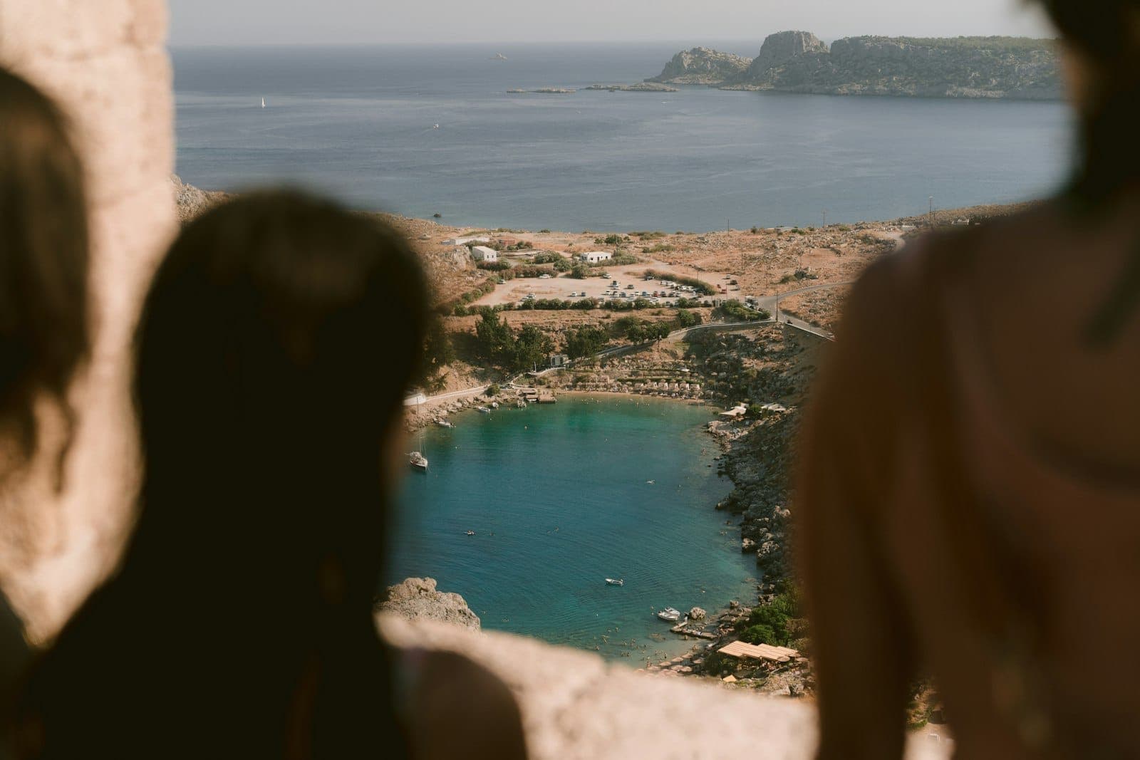 Vue aérienne de la baie de Lindos à Rhodes avec des eaux turquoise, des parasols sur la plage, un littoral rocheux et des caps au loin.
