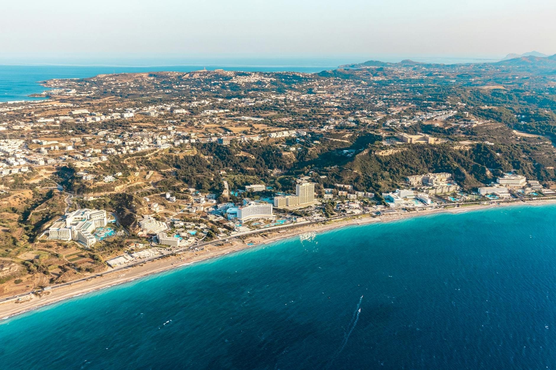 Aerial view of Rhodes coastline with hotels, town buildings, and deep blue sea under clear sky