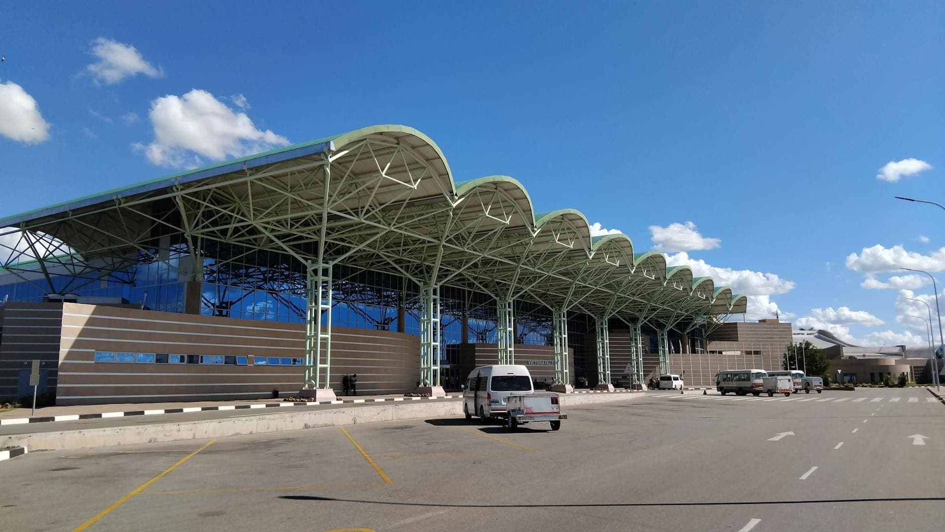 Exterior view of Rhodes International Airport terminal with distinctive wavy roof, parked vehicles, and clear blue sky.