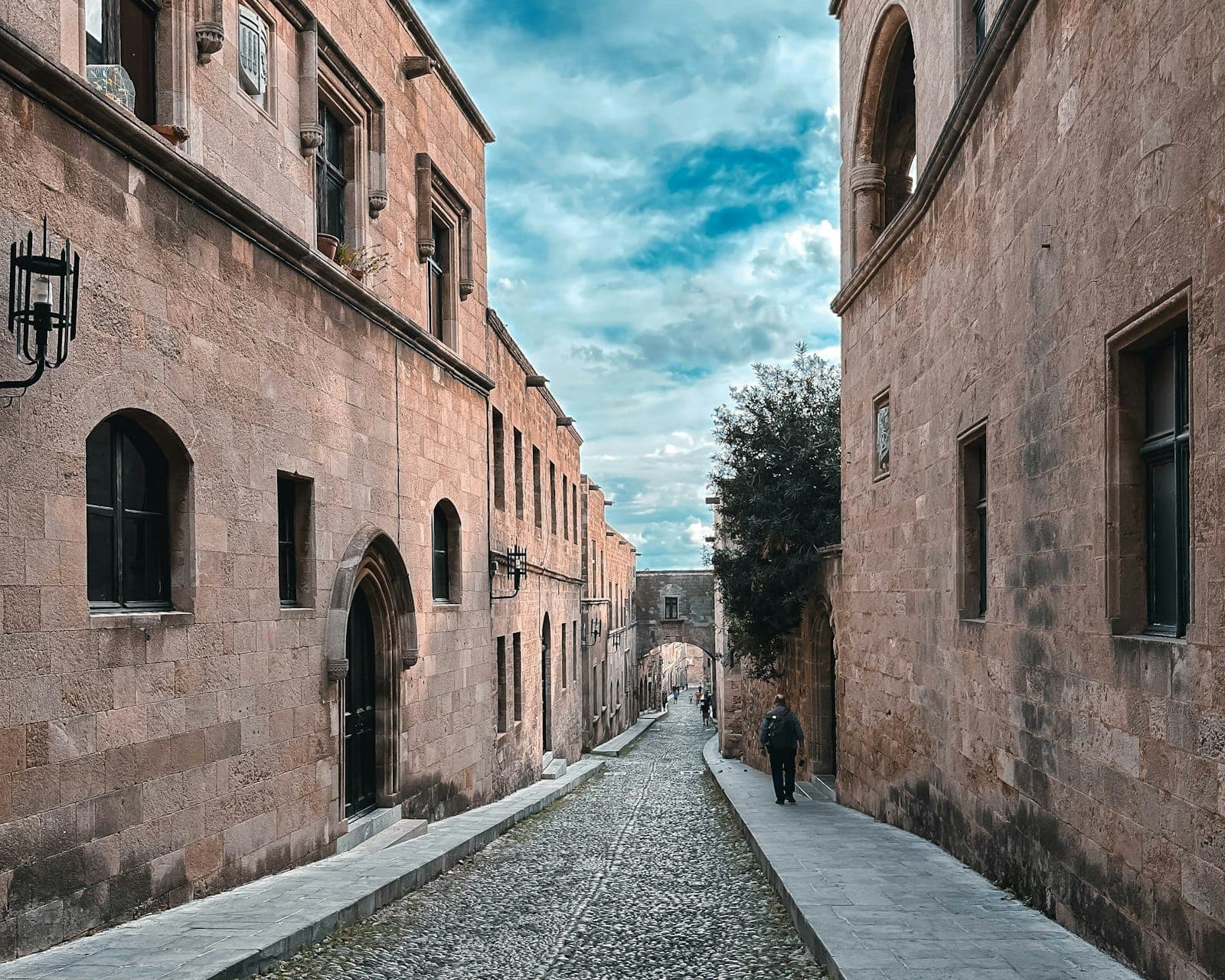 A wide cobbled street lined with medieval stone buildings in Rhodes Old Town, leading to an arched gateway under a blue sky.